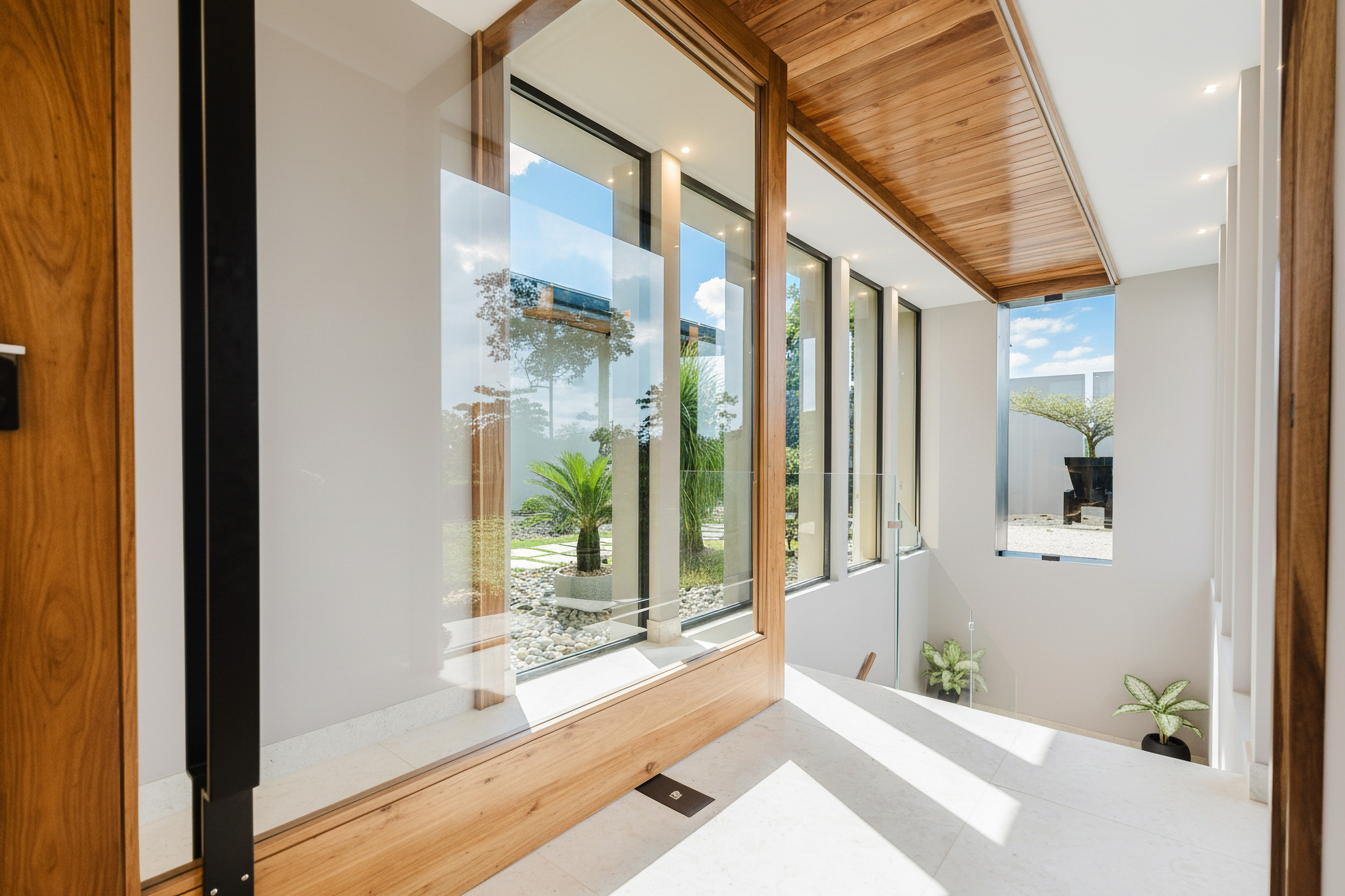 This bright interior hallway features expansive glass walls with warm wood framing, offering a tranquil garden view and natural light spilling over the light-toned floors towards a descending staircase.