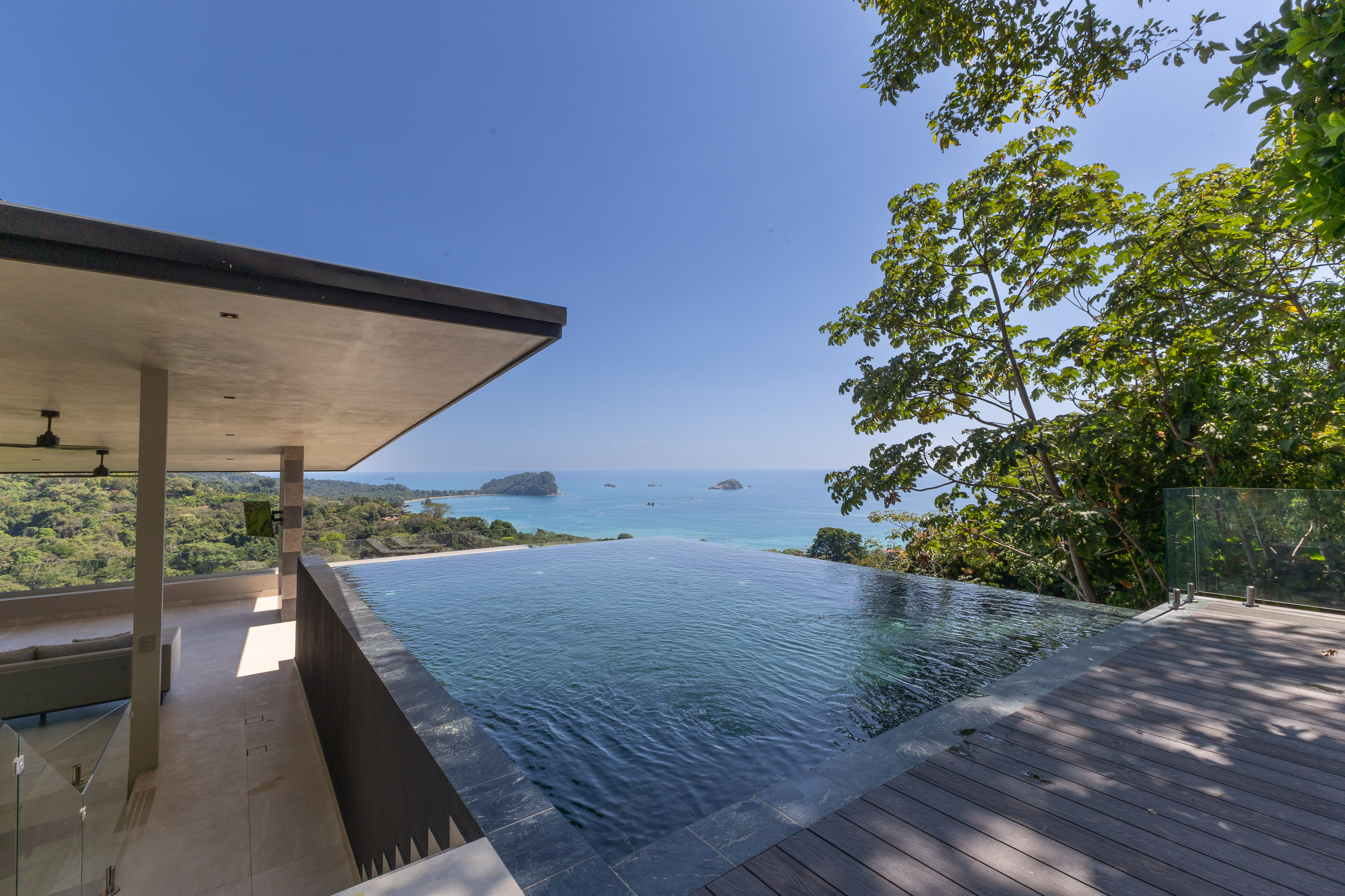 A rooftop infinity pool with a dark tiled edge overlooks the Pacific Ocean and a verdant coastline under a clear blue sky, adjacent to a covered outdoor lounge area.