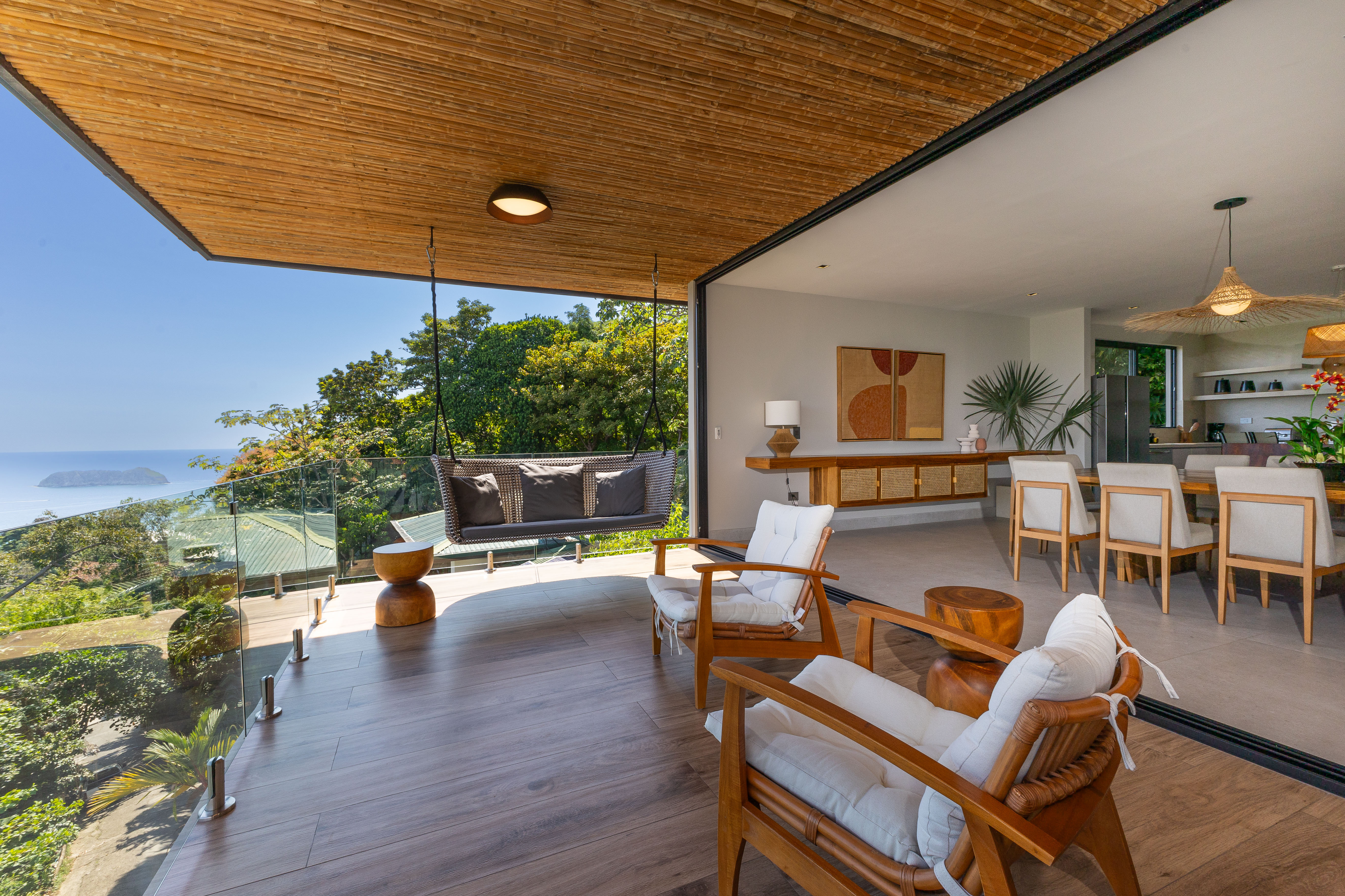 A contemporary open-plan terrace features a slatted wood ceiling, comfortable lounge seating, and glass railings framing ocean and jungle views, merging with an indoor dining area.