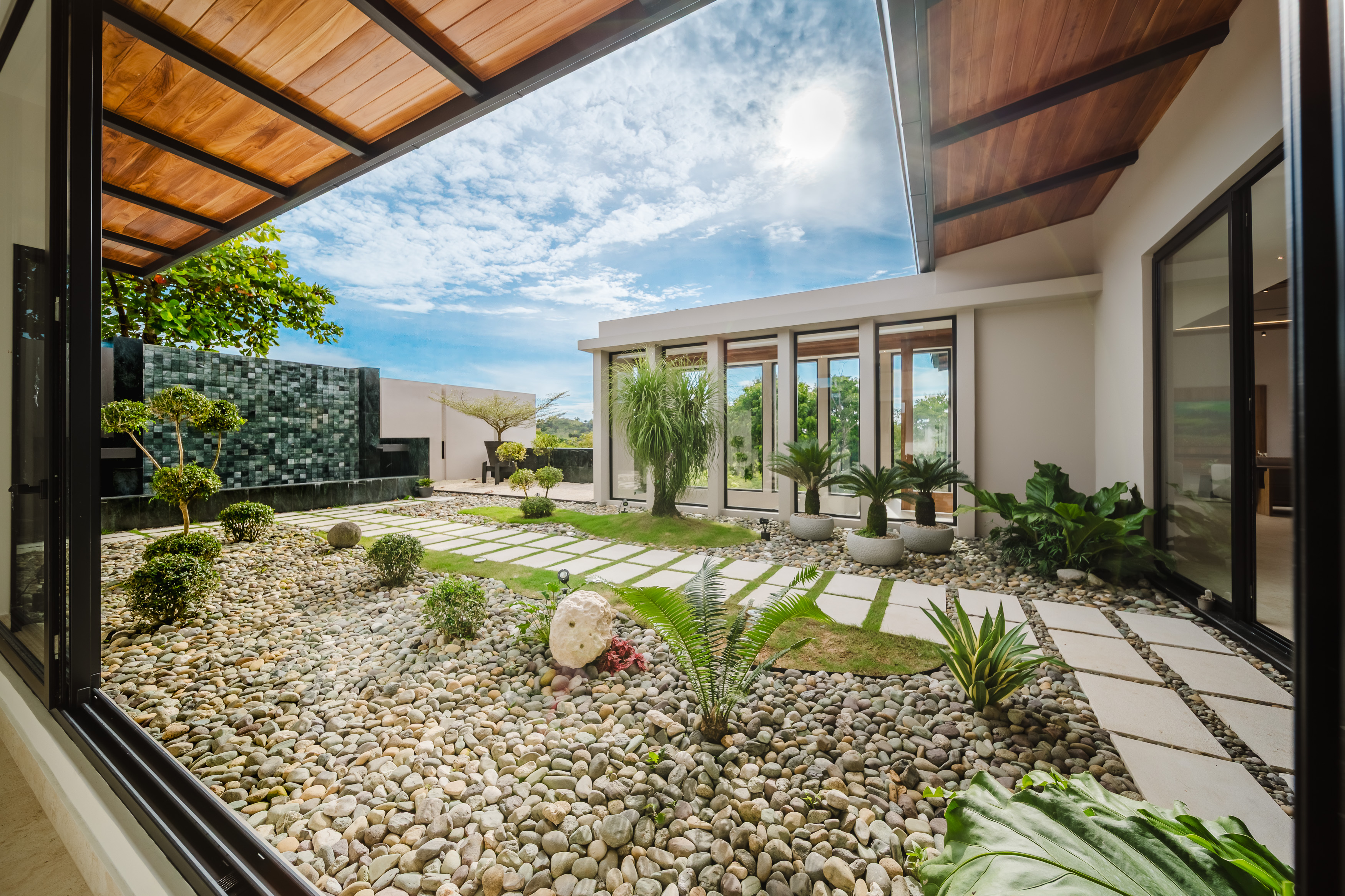 Large open sliding glass doors reveal a sunlit private garden with pebble ground, geometric stepping stones, diverse tropical plants, and a modern water feature, framed by a timber ceiling and contemporary architecture.