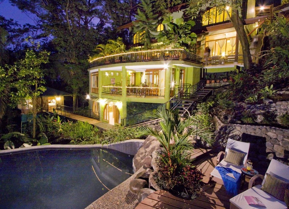 An illuminated multi-level villa is nestled into a dense jungle landscape, featuring open-air balconies, an outdoor pool, and a secluded poolside deck.