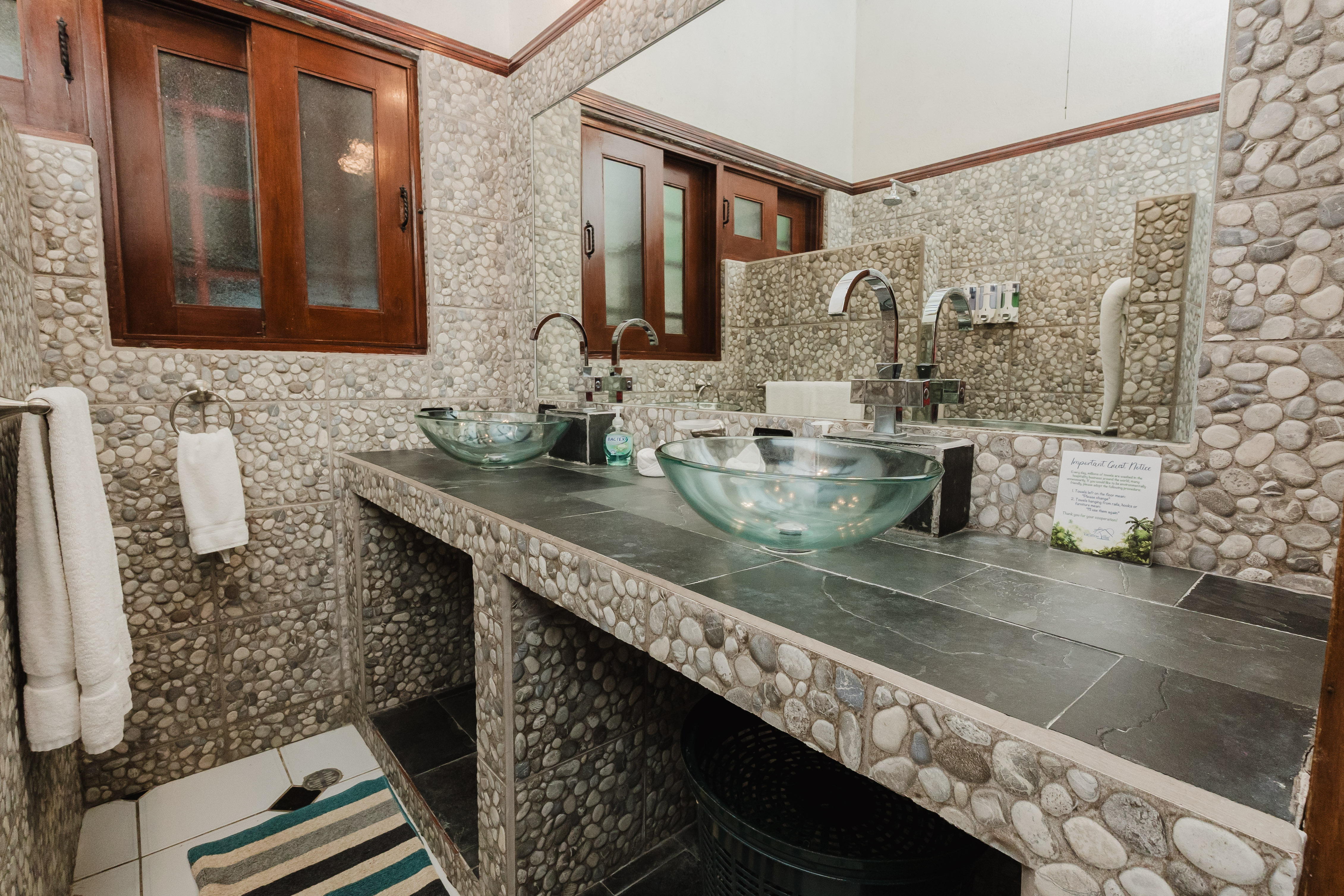 The bathroom features pebble-tiled walls, a dark stone vanity with two glass vessel sinks, and a wooden-framed window allowing natural light.