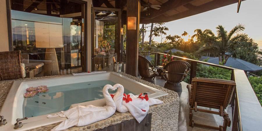 An outdoor jacuzzi is set on a stone-edged balcony with seating, providing tropical forest views under a warm sunset glow.