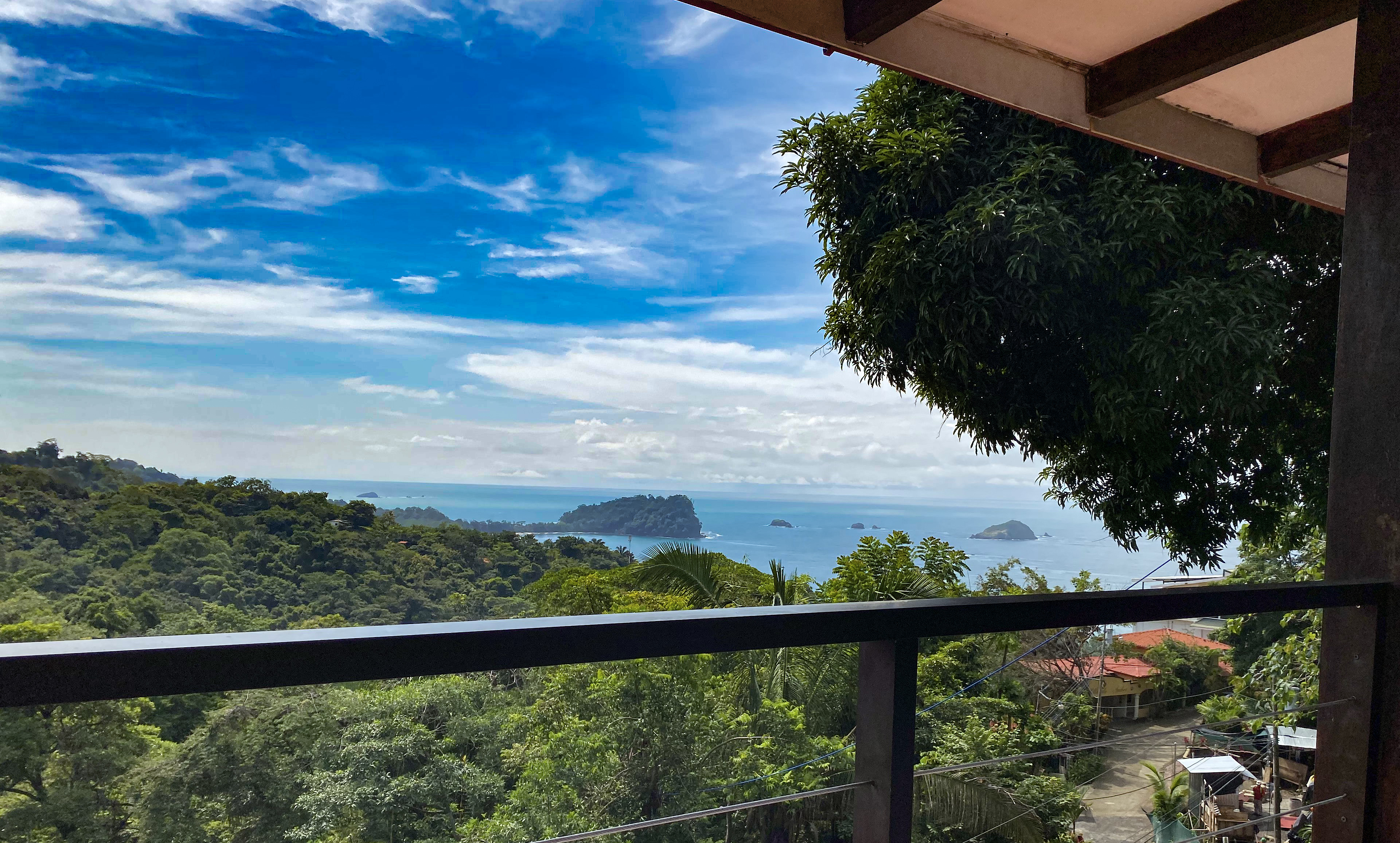 From a shaded terrace, a wide view extends over lush tropical jungle to the Pacific Ocean, revealing several small islands and a bright, cloud-streaked sky.