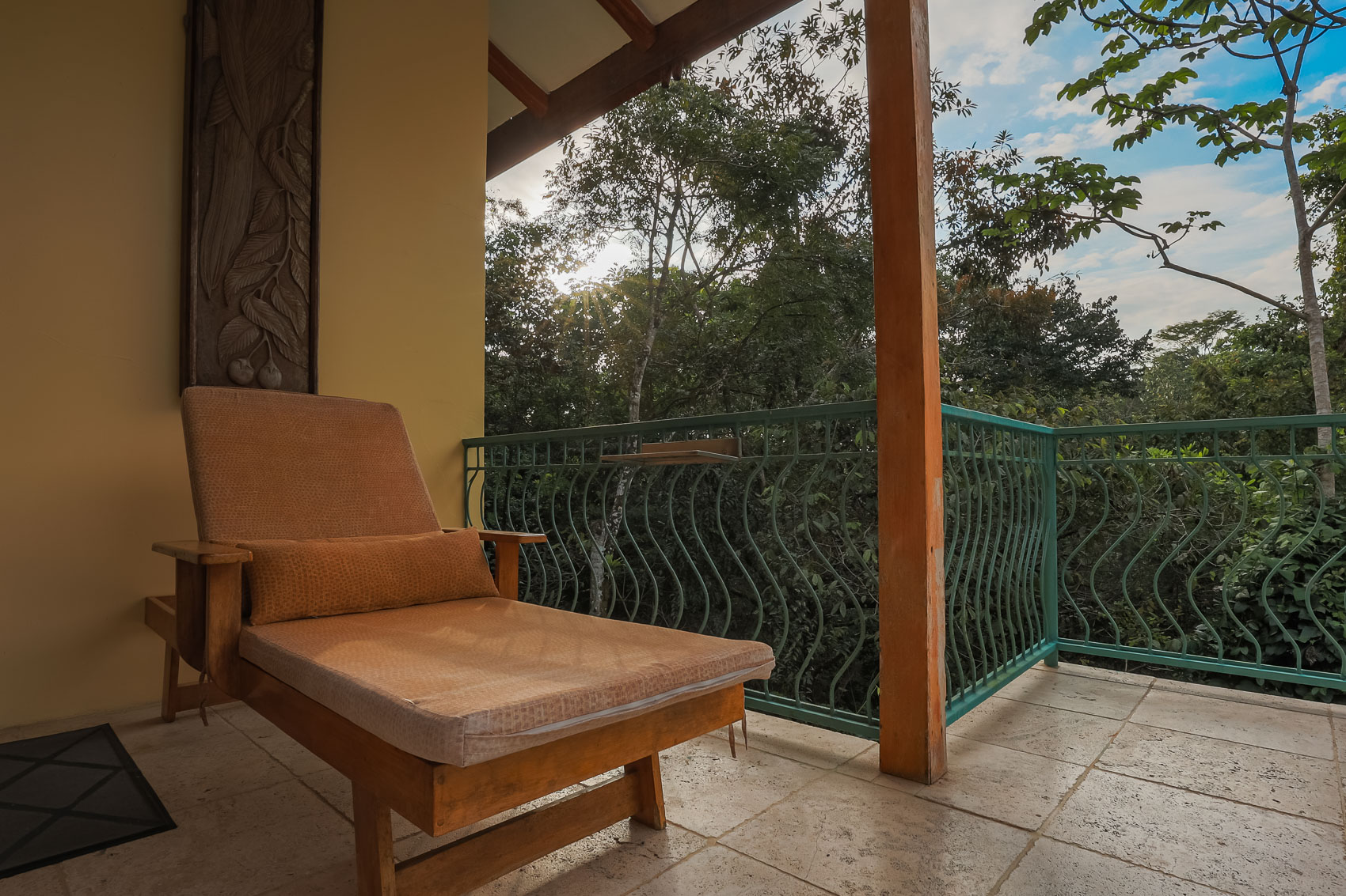 A private covered balcony, featuring a carved wall panel and a comfortable chaise lounge, opens to immersive views of the dense tropical canopy.