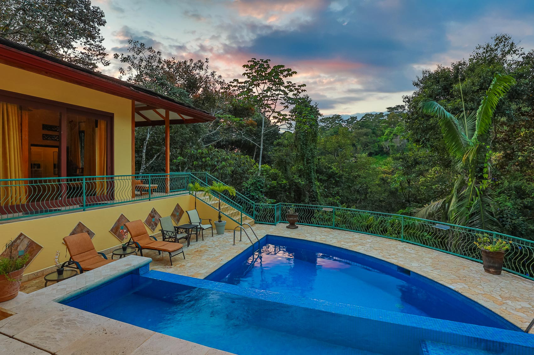 A two-tiered swimming pool and stone terrace are visible at dusk, bordered by a yellow villa and dense tropical forest.