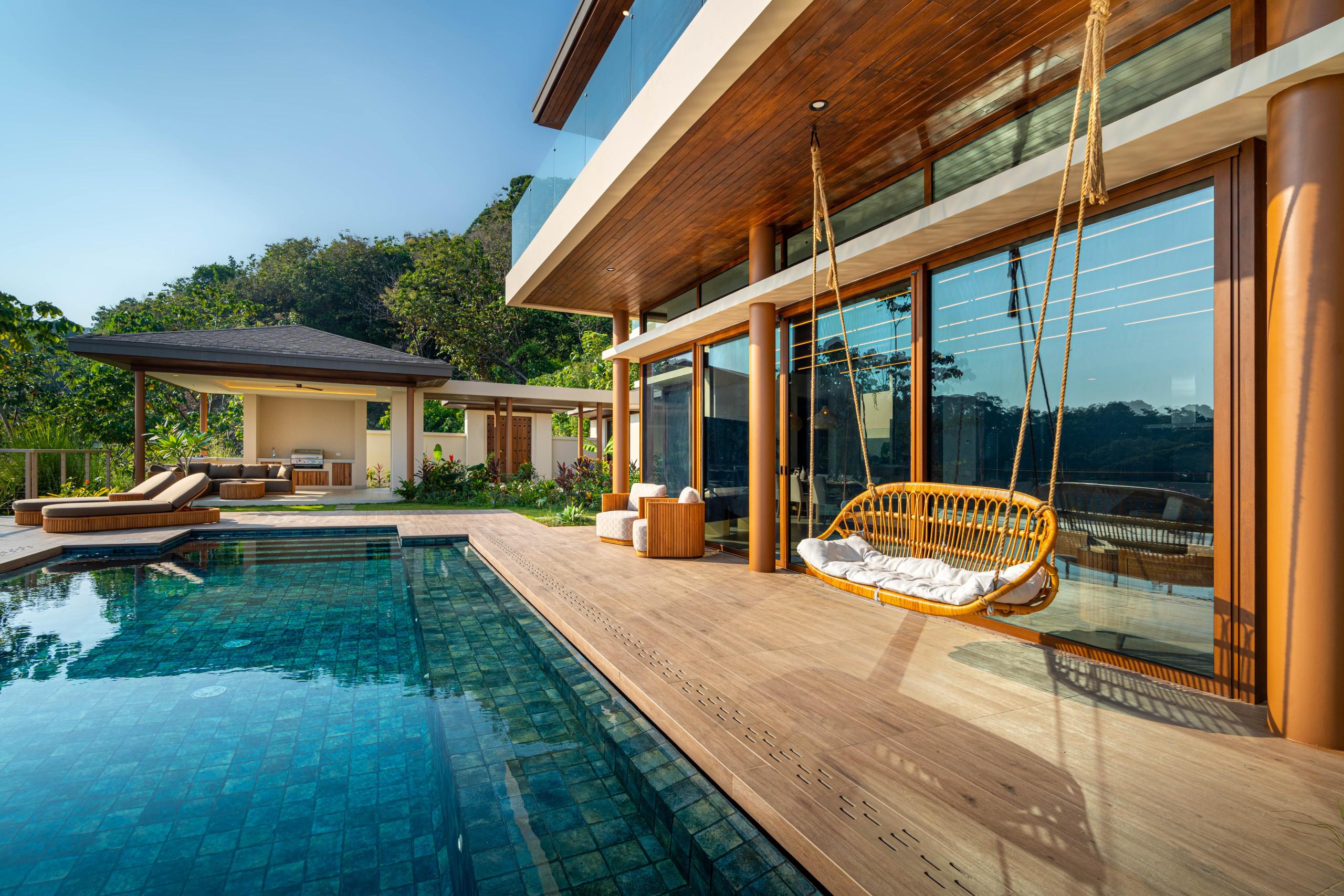 A deep blue tiled infinity pool extends from a light wooden deck, fronting a modern villa with full-height glass doors and views of the surrounding tropical jungle under a clear sky.