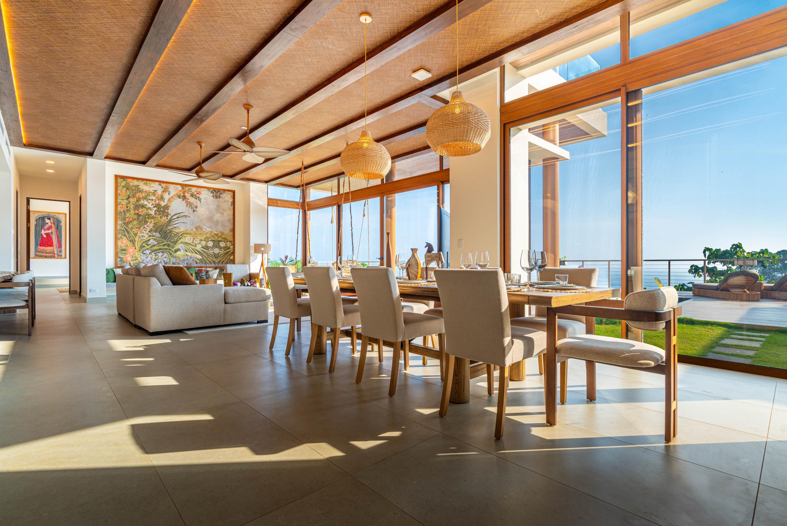 This spacious living room, featuring a textured woven ceiling and exposed wooden beams, is bathed in natural light from floor-to-ceiling windows that reveal expansive views of the sky and outdoor deck.