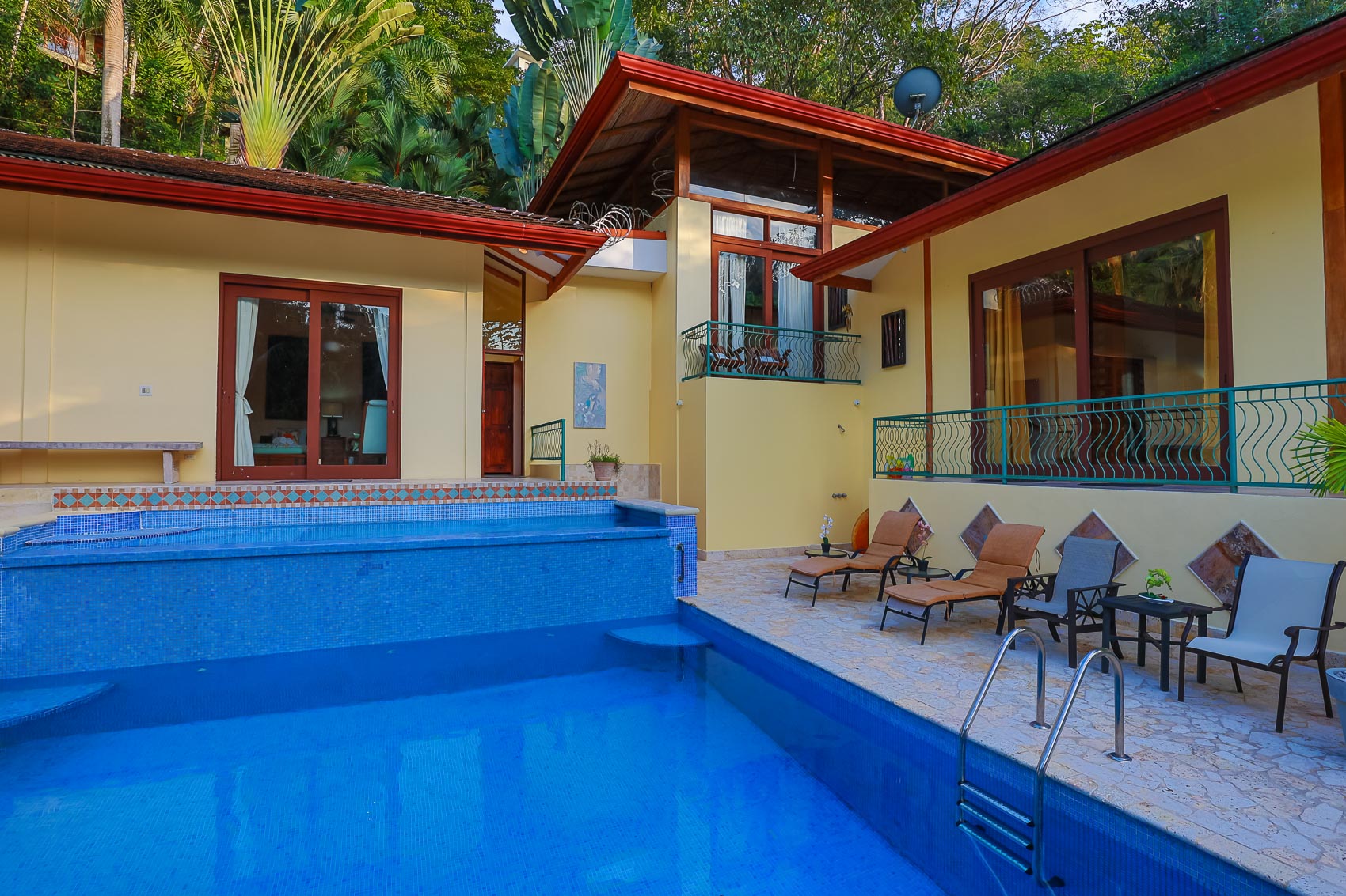 The villa's yellow exterior, featuring distinctive red-tiled roofs and multiple balconies, overlooks a two-tier blue mosaic pool and a spacious stone patio surrounded by vibrant tropical foliage.