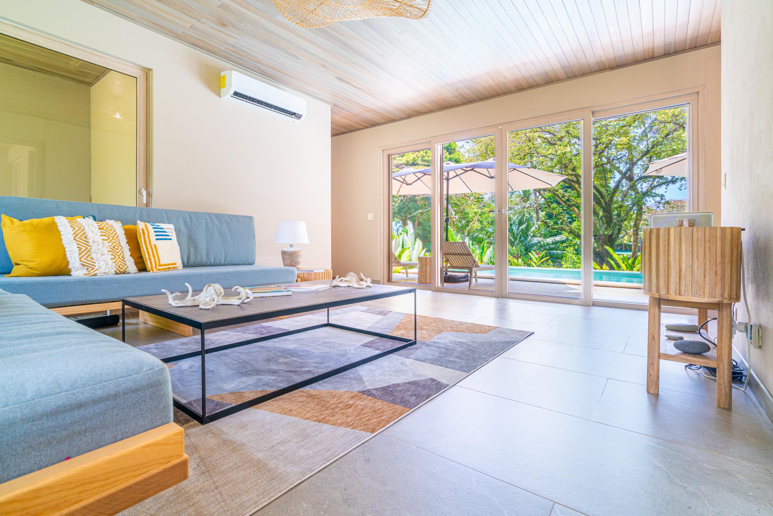 This living room features a wood-paneled ceiling and large glass doors that open directly onto a private pool and verdant jungle, allowing abundant natural light.