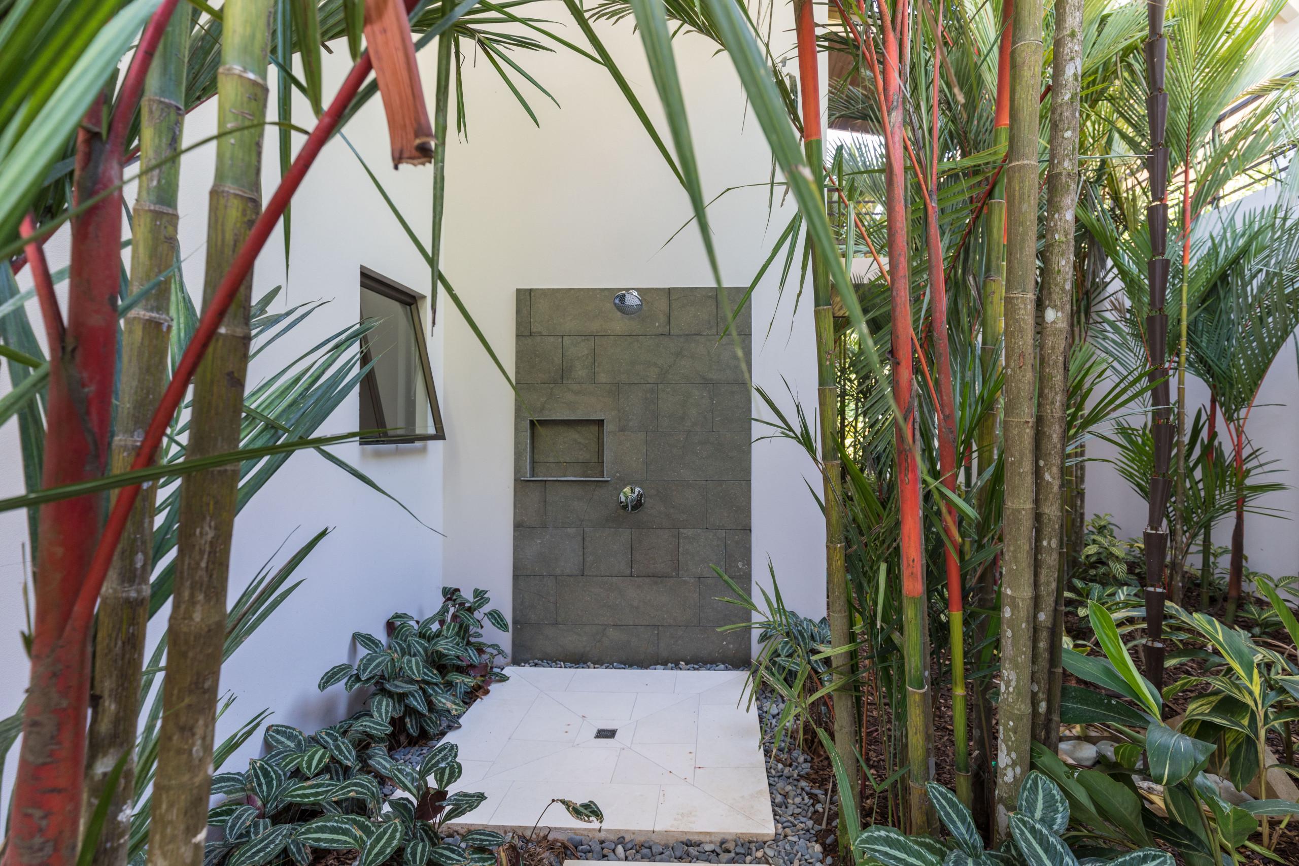 A private outdoor shower features a dark stone-tiled wall with a shower head, surrounded by lush tropical plants and receiving ample natural light.