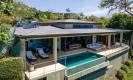 The contemporary villa features an infinity-edge pool extending from a spacious, covered outdoor terrace, positioned on a hillside overlooking the surrounding landscape.