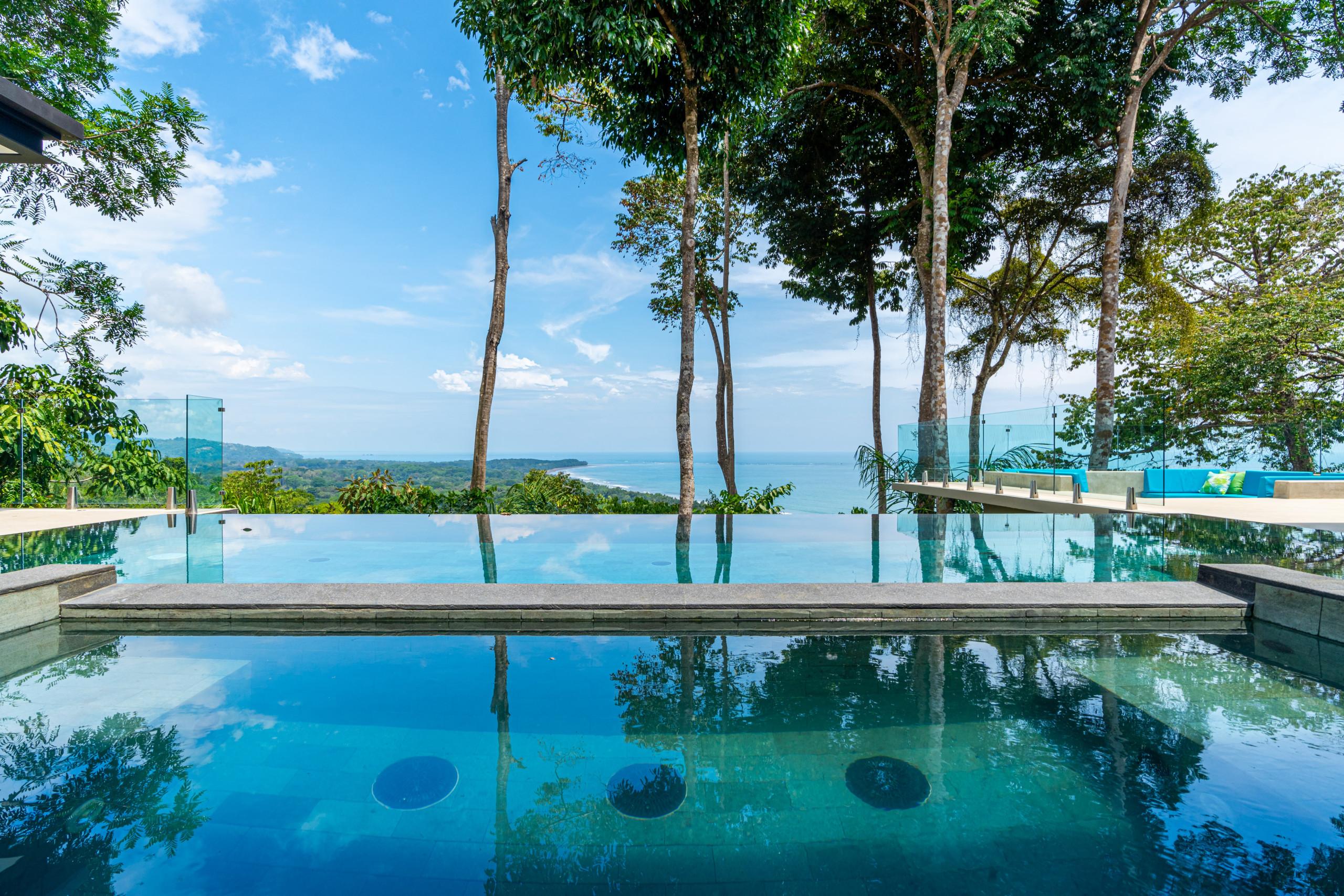An expansive infinity pool offers uninterrupted ocean and coastline views under a bright blue sky, framed by tropical trees and transparent glass railings.