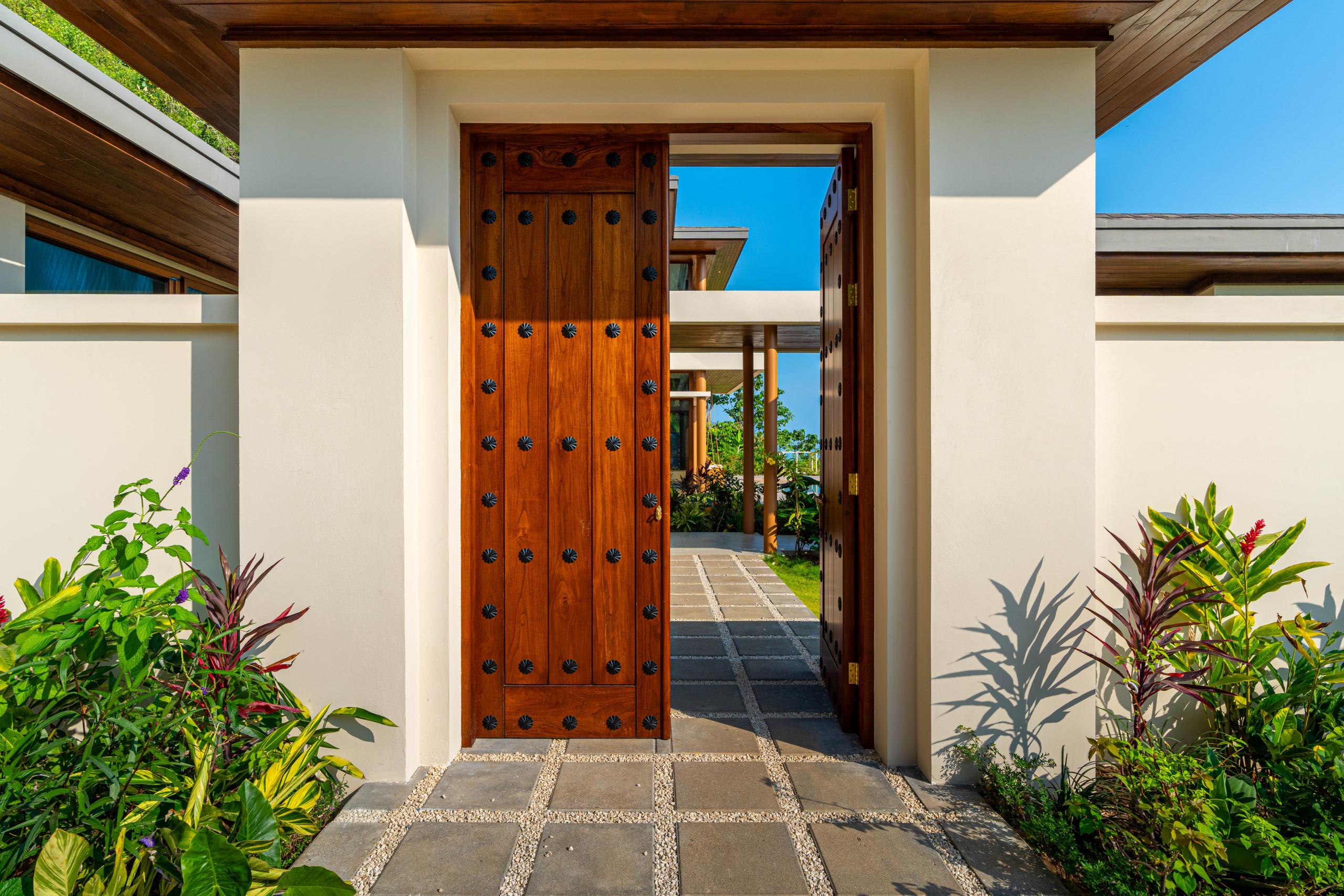 The studded wooden entrance door, part of the Torana-style architecture, stands open to a sunlit stone path lined with tropical plants, leading into an open courtyard.
