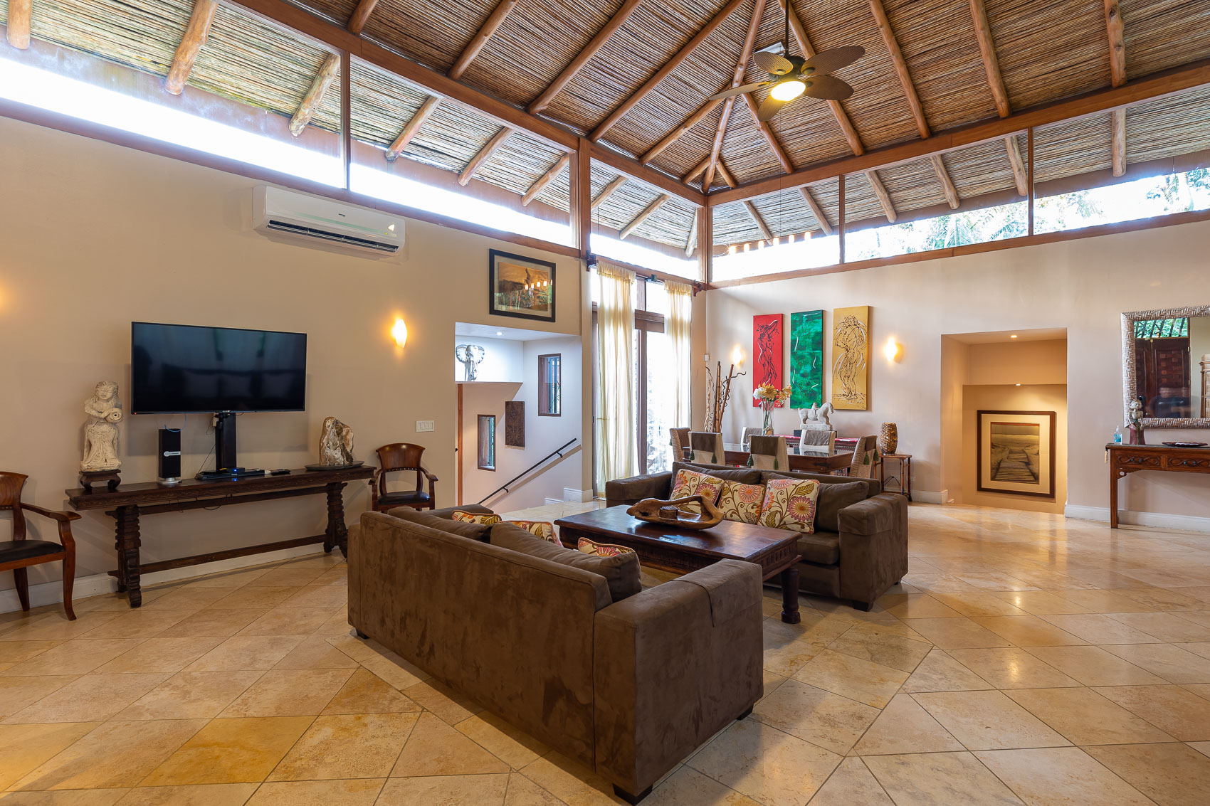A spacious open-concept living and dining area is illuminated by natural light from clerestory windows and features a high exposed wooden beam and bamboo ceiling over tiled floors.