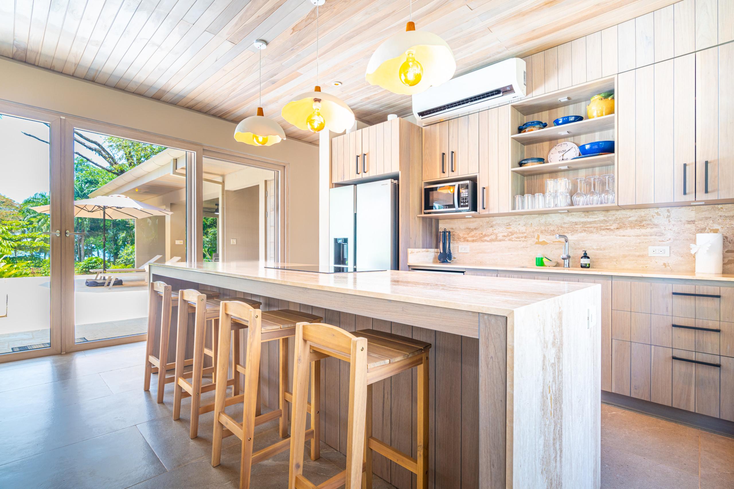 The kitchen features light wood cabinetry, a stone island with integrated seating, and large glass doors opening to an outdoor terrace with views of the lush jungle and distant ocean.