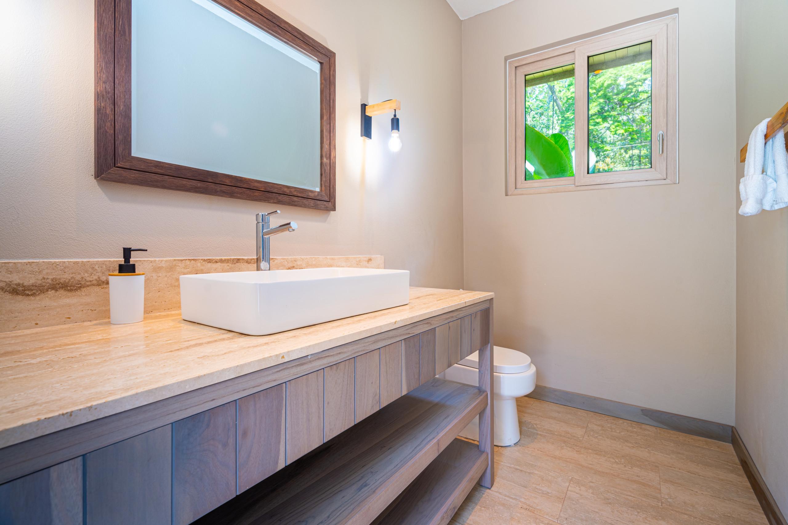 This bathroom features a natural wood vanity with a stone countertop, a modern vessel sink, a framed mirror, and a window providing a view of the surrounding lush jungle.