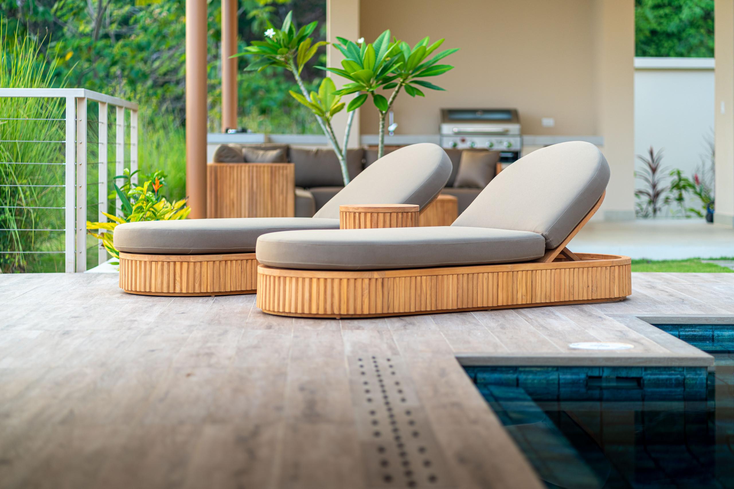 A wooden deck with two sun loungers adjoins an infinity pool, with a covered outdoor kitchen and sofa visible among tropical foliage.