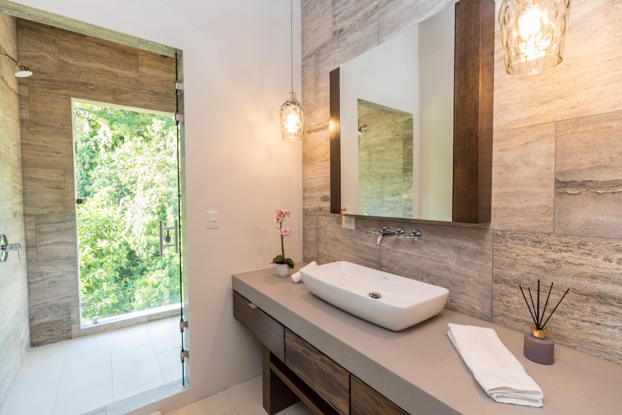 This modern bathroom features a floating vanity with a vessel sink and wall-mounted faucet, stone-tiled walls, and a glass-enclosed shower with a direct opening to a lush jungle view.