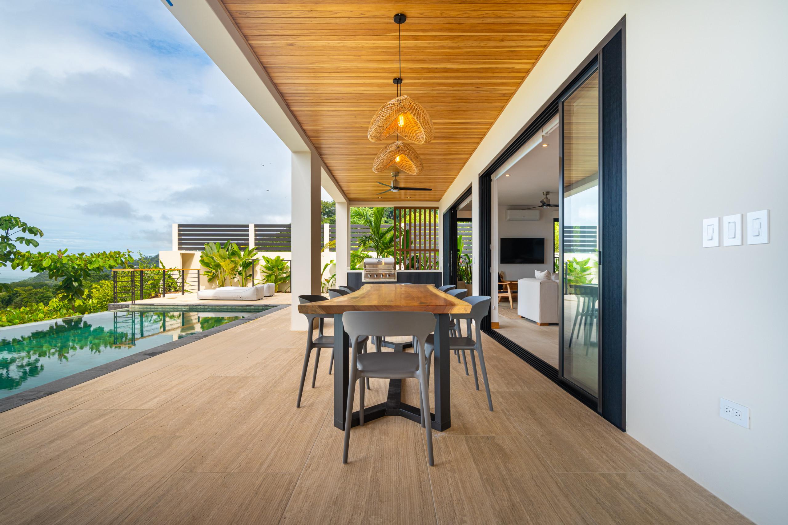 This spacious covered outdoor terrace features a long dining table, an infinity pool, and expansive views of lush greenery and the ocean under a bright sky, providing a seamless transition to the interior living space.