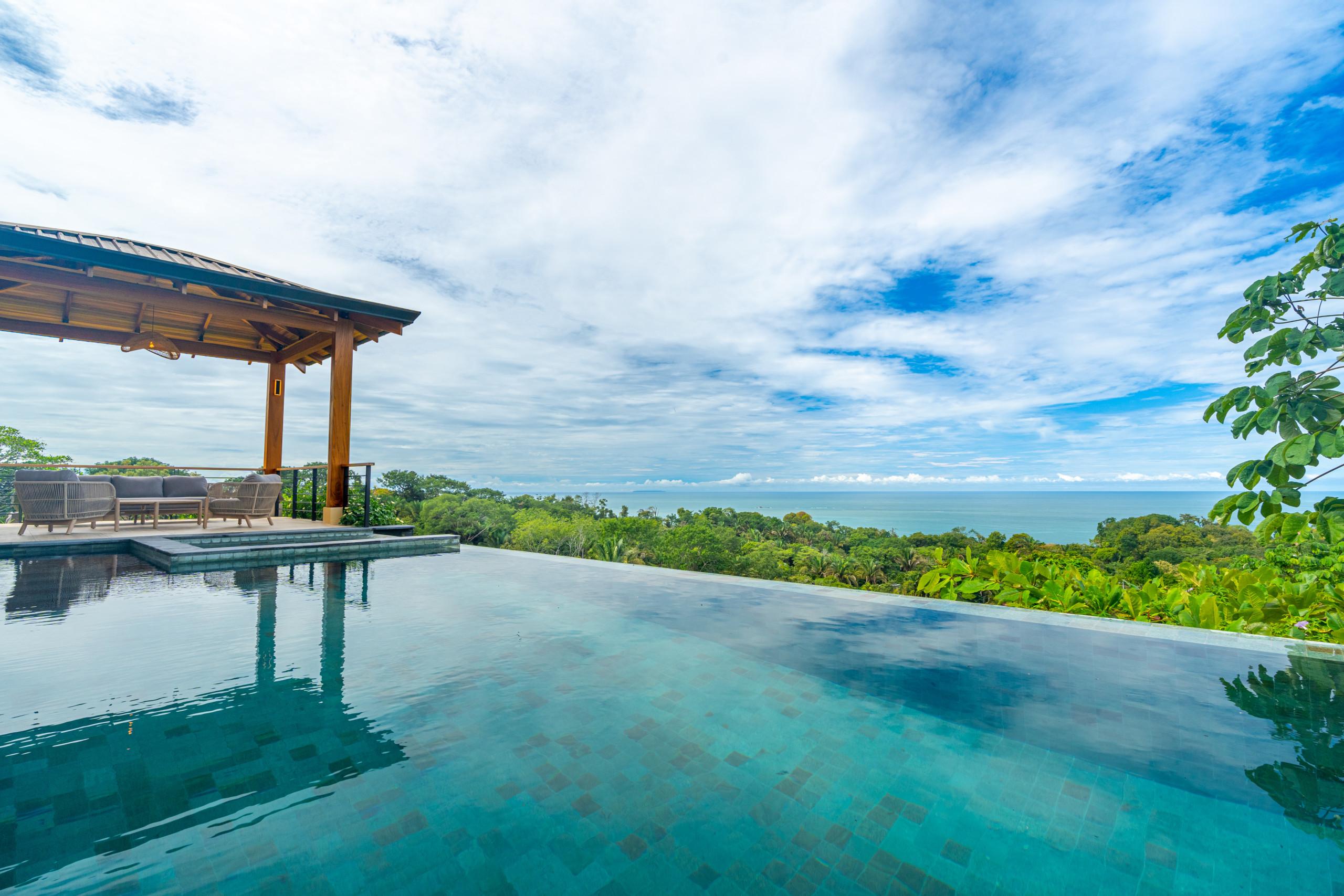 An infinity pool with a shaded palapa lounge area extends toward the Pacific Ocean and lush jungle under an open sky.