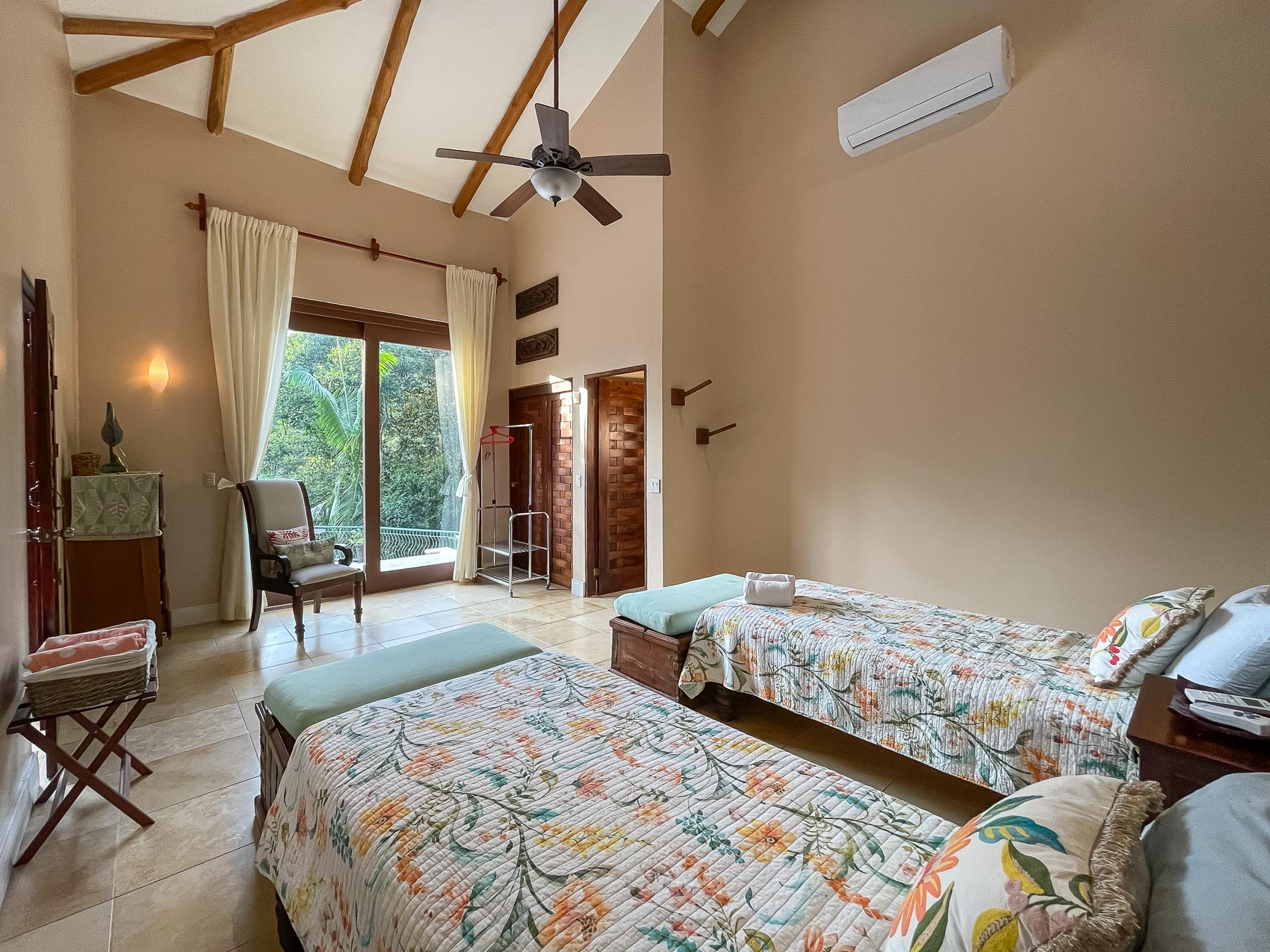 This spacious bedroom features two individual beds, a vaulted ceiling with exposed wooden beams, and glass sliding doors that open to a lush jungle view, creating an open and bright atmosphere.
