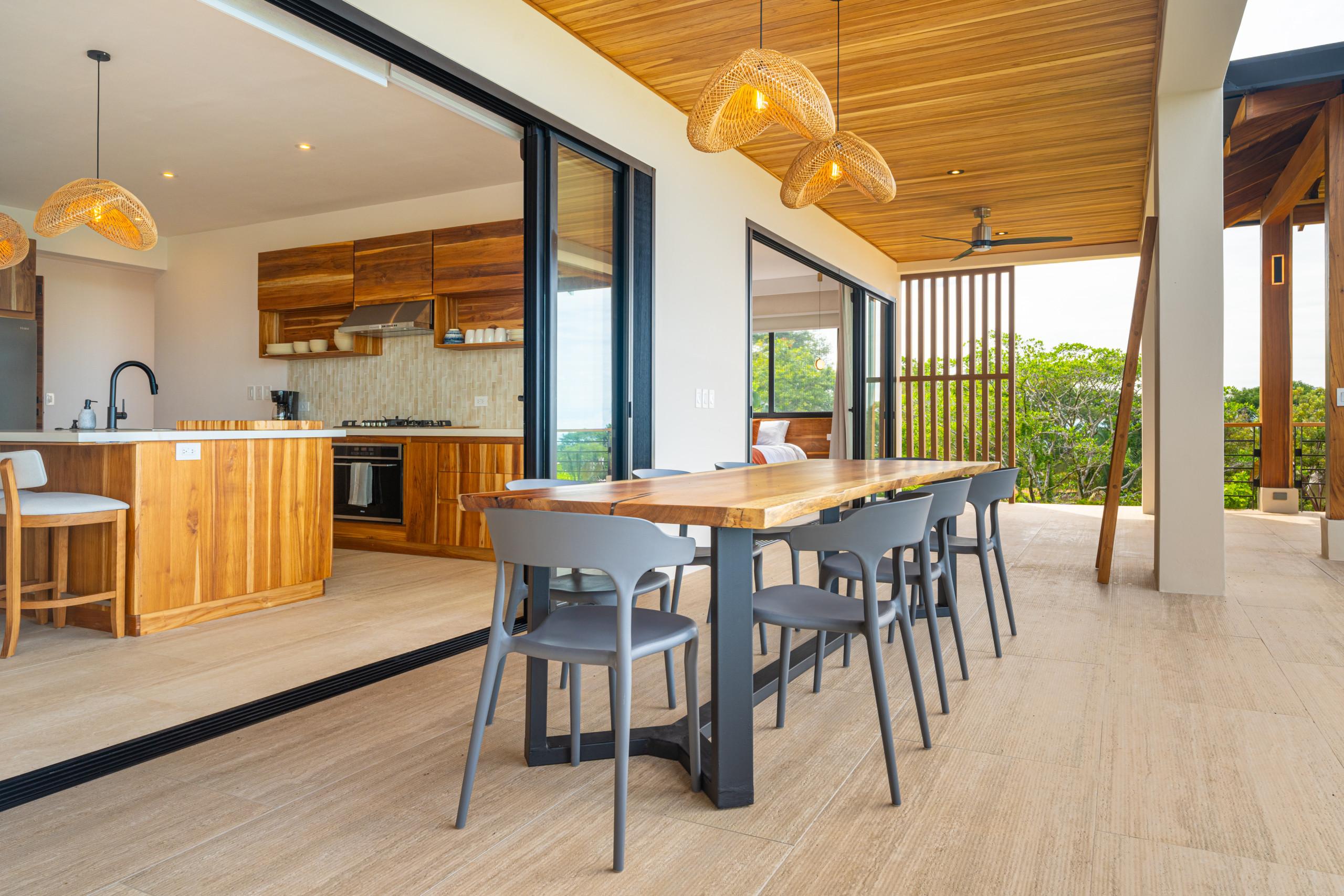 An outdoor terrace features a wooden dining table and grey chairs under a natural wood ceiling, opening to an indoor kitchen via sliding glass doors and offering views of surrounding trees.