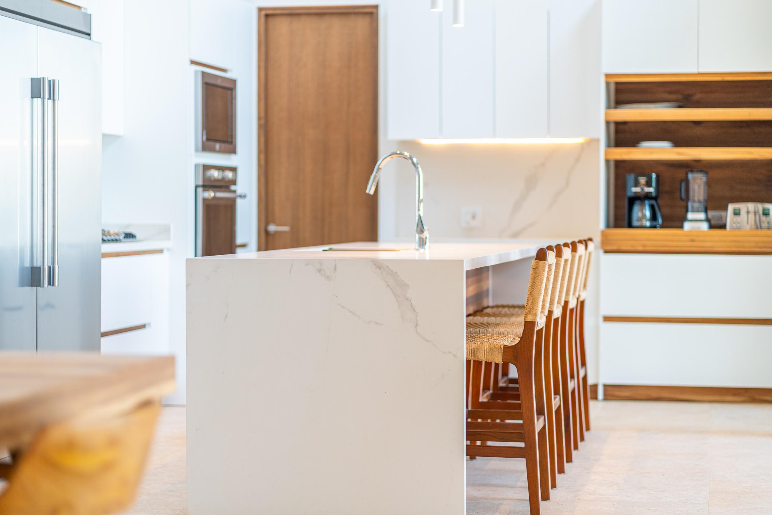A bright, contemporary kitchen features a long island with an integrated sink and bar stools, alongside white cabinetry, built-in appliances, and open wood shelving.
