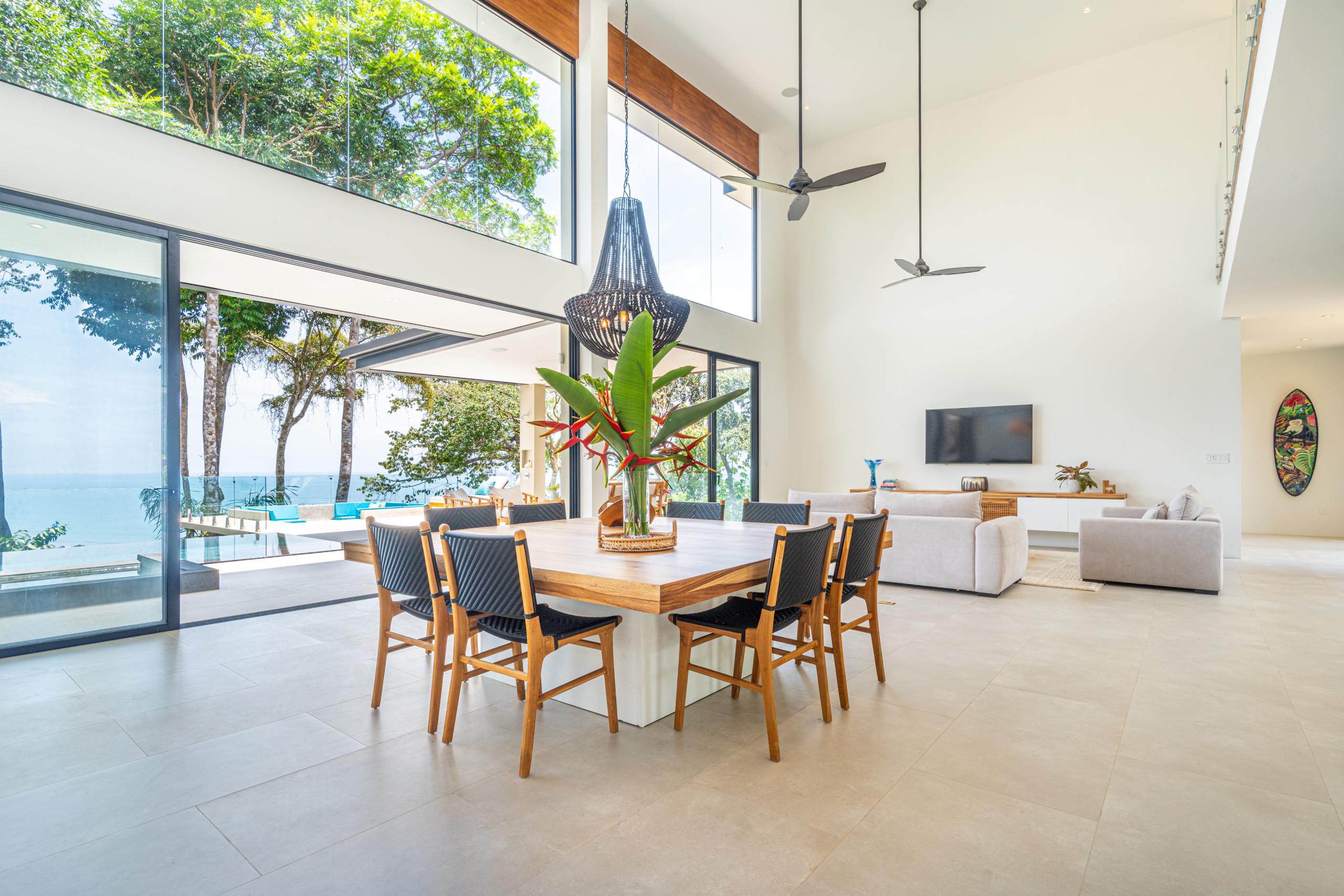 The double-height dining area is filled with natural light from floor-to-ceiling glass doors and windows, offering seamless ocean views framed by tropical foliage.