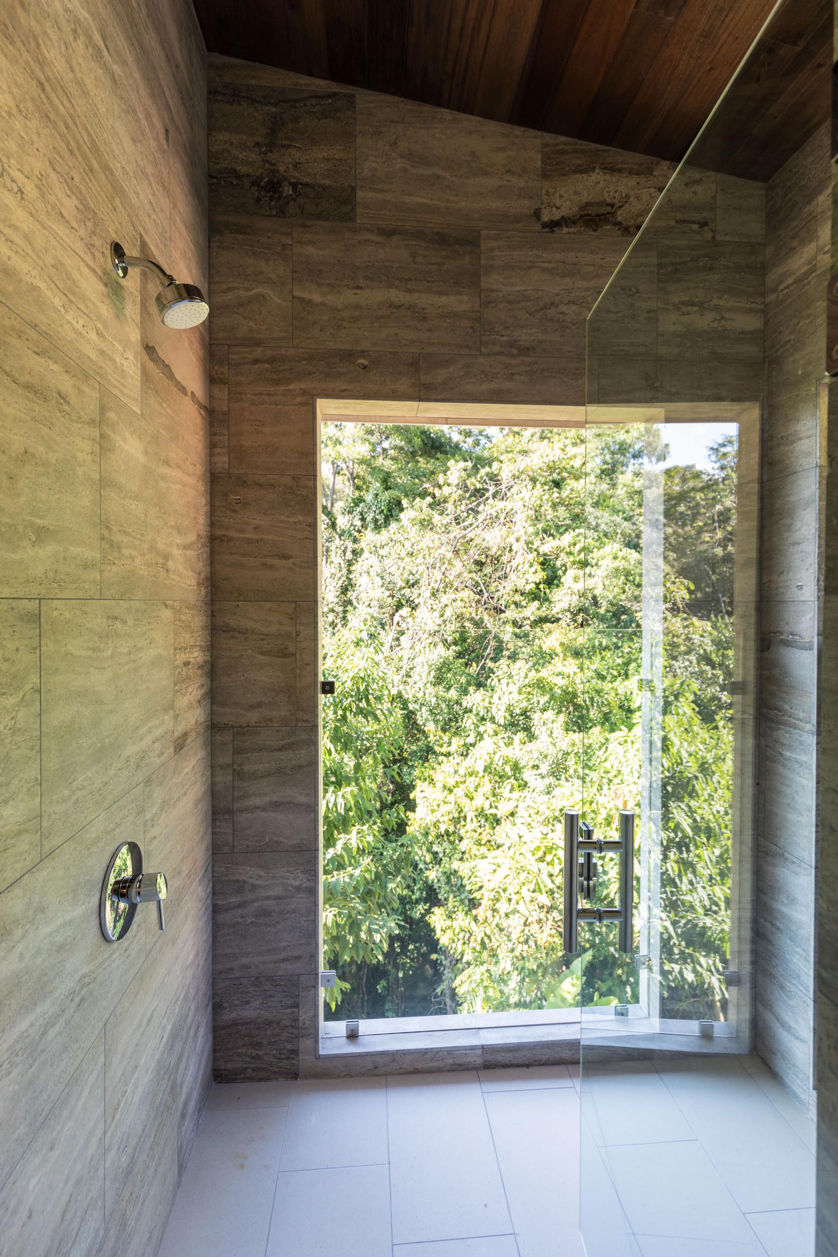 A stone-tiled shower with a clear glass enclosure features a large window revealing lush jungle foliage, bringing abundant natural light and views into the space.