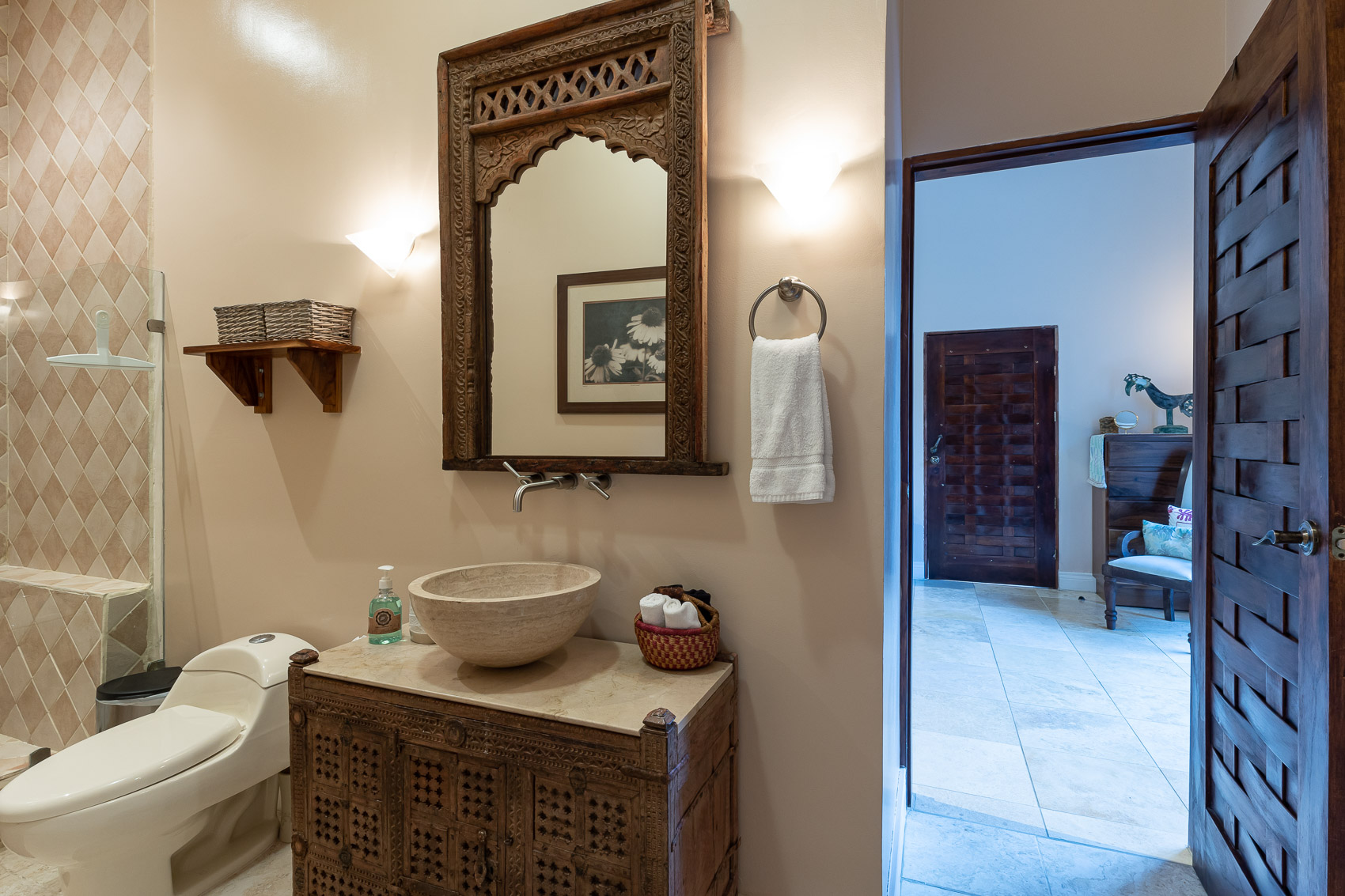 This Balinese-inspired bathroom features a stone vessel sink set on a carved wooden vanity with a large matching mirror, alongside a tiled shower, and an open doorway leading to another area of the villa.