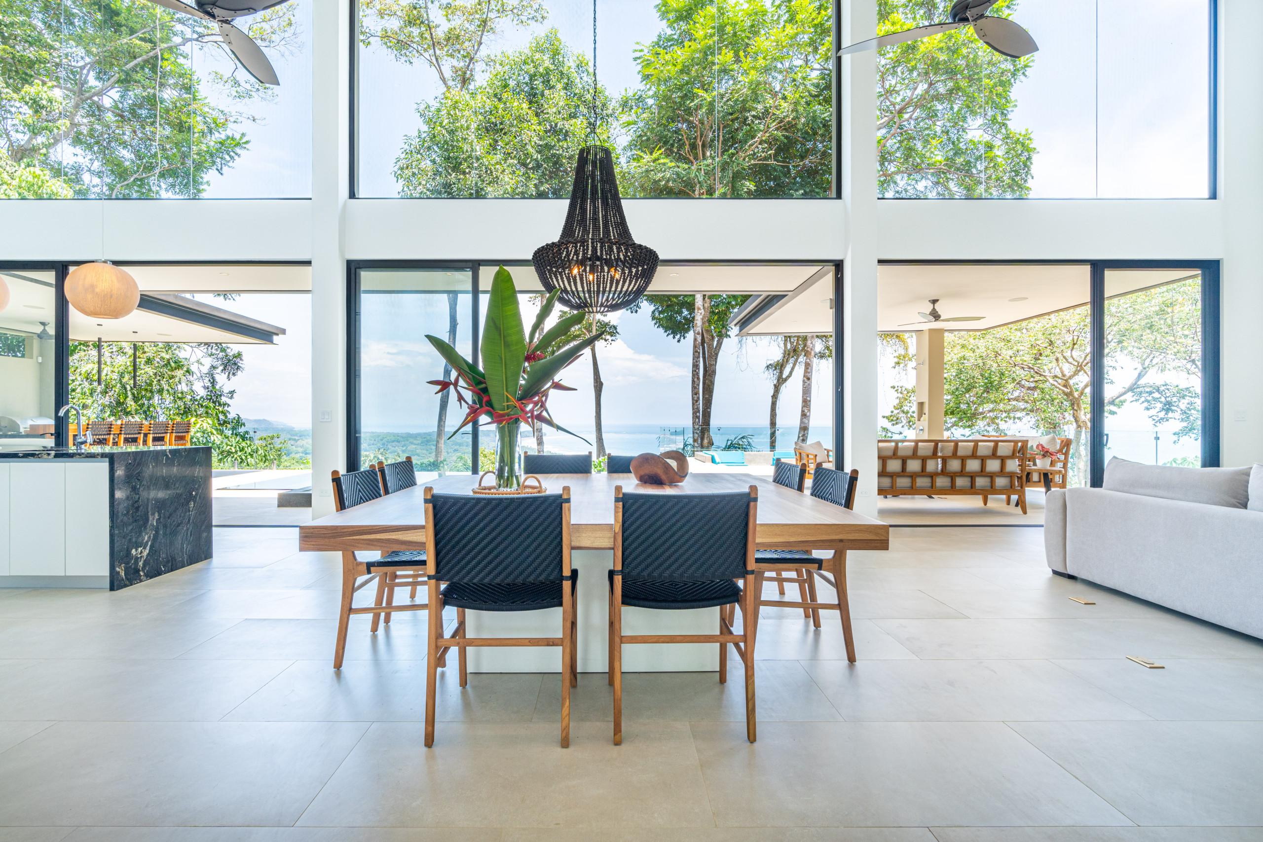 This bright, open-concept living space integrates a dining area and features floor-to-ceiling glass walls showcasing panoramic ocean and jungle views.