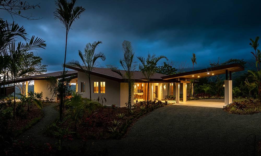 The modern villa, with its angled rooflines and warmly lit interior, is nestled amidst lush tropical landscaping and palm trees, featuring a covered entry and carport at twilight.