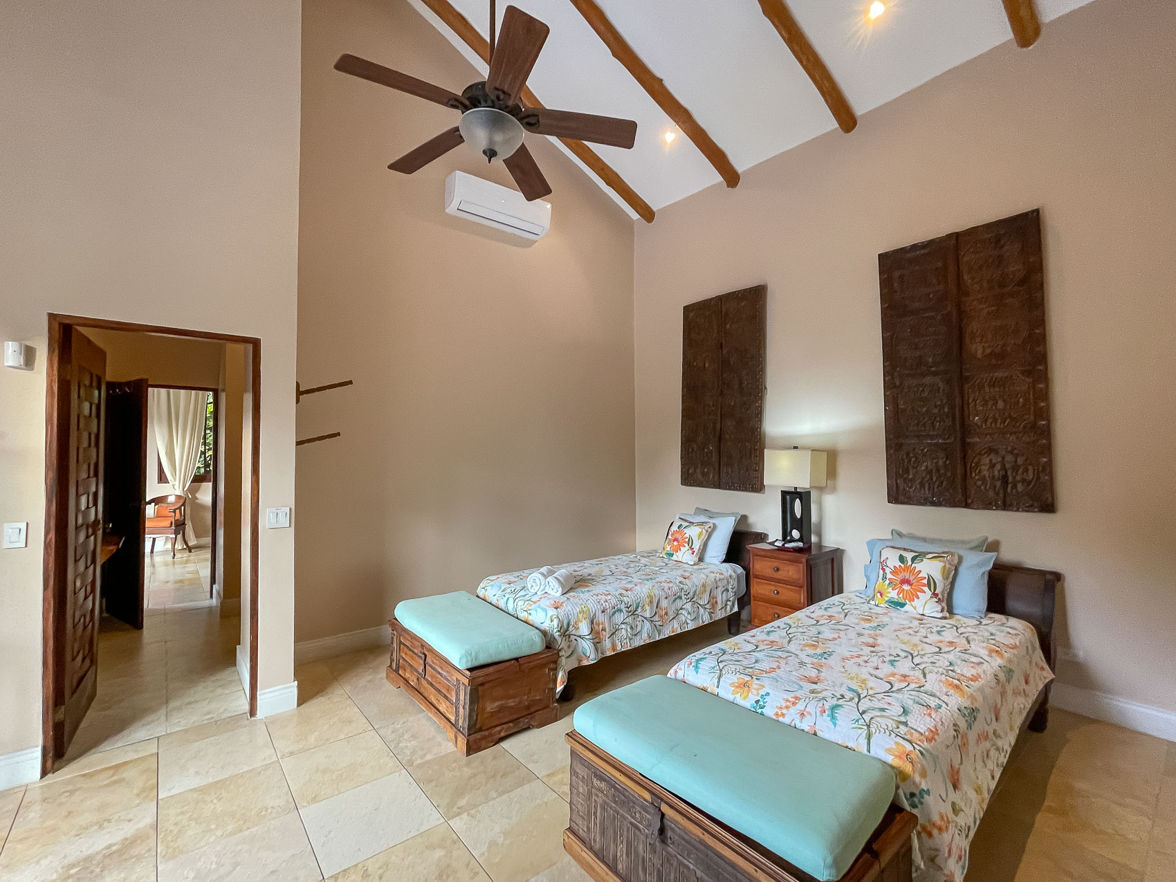This bedroom offers a high vaulted ceiling with exposed wooden beams, contains two individual beds, and is styled with Balinese-inspired wooden details.