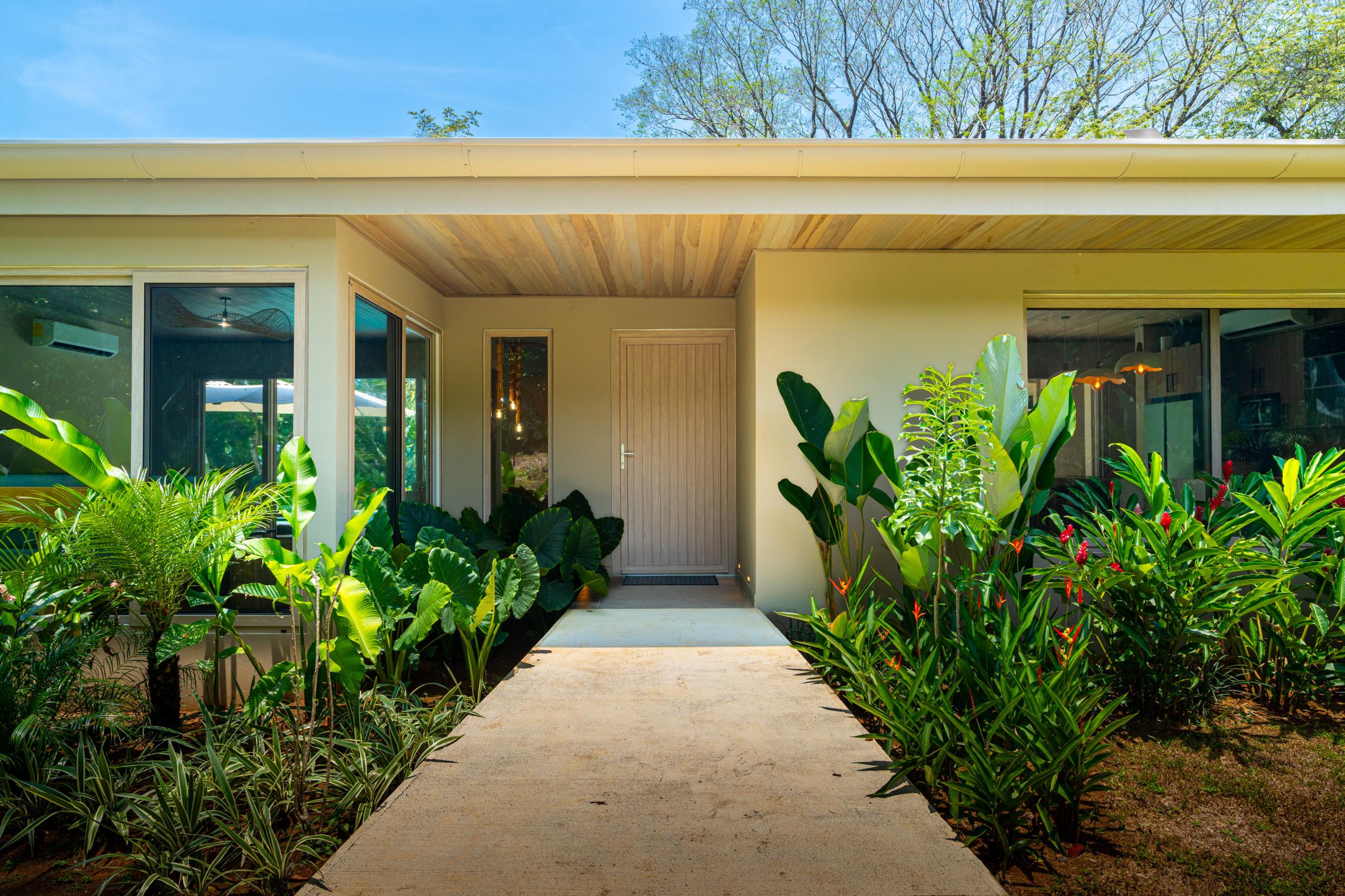 An inviting concrete pathway leads to the villa's main entrance, featuring a warm wooden door, planked ceiling, large windows, and vibrant tropical landscaping bathed in natural light.