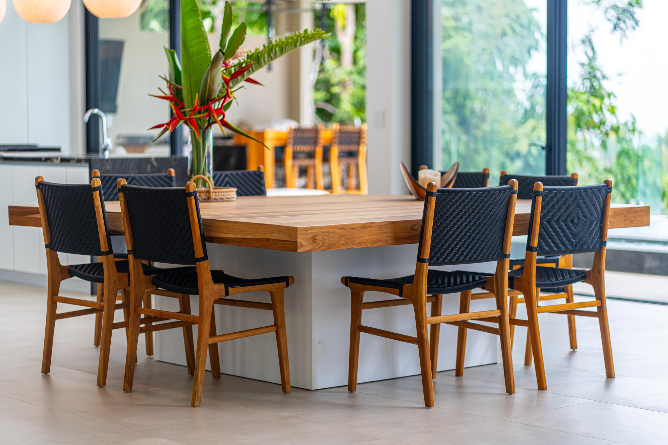 This open dining area features a substantial wooden table and woven chairs, flooded with natural light from expansive windows that frame tropical views.