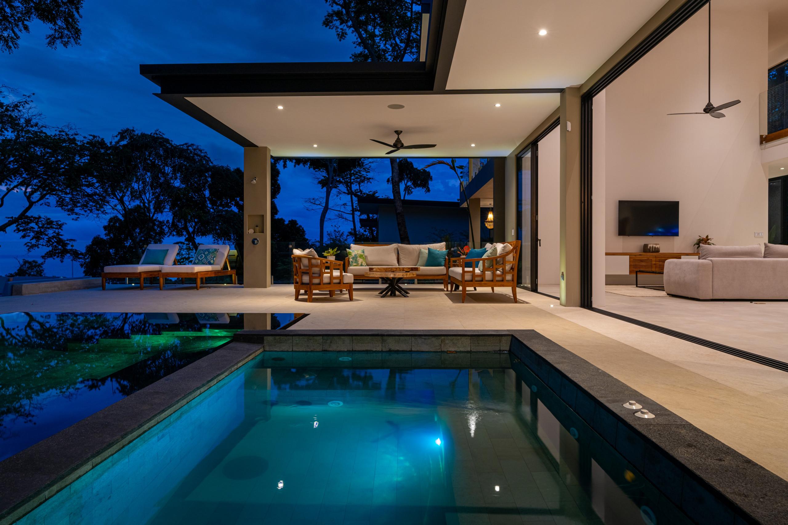 An inviting illuminated infinity pool and adjacent jacuzzi are set before a covered outdoor lounge area with contemporary seating, which opens through expansive glass doors to an interior living space, all under the serene twilight.
