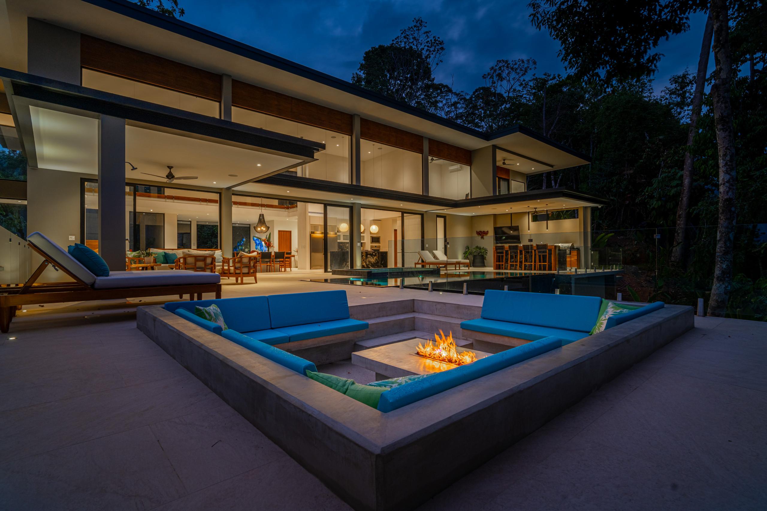 The modern villa's illuminated exterior features an expansive outdoor living space with a sunken fire pit, surrounding lounge seating, an infinity pool, and an outdoor kitchen and bar, set against tropical forest at night.