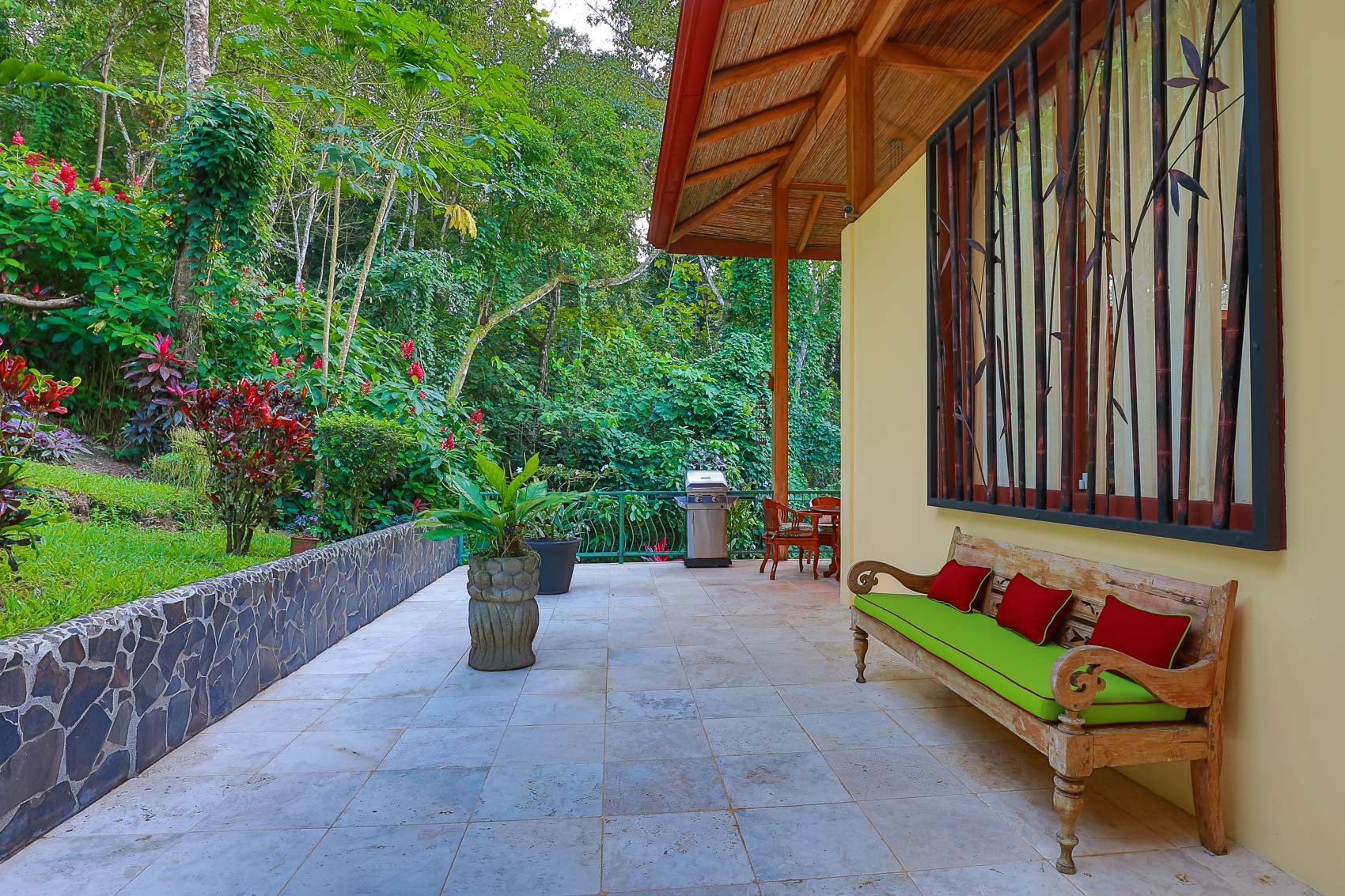 A private outdoor terrace with tiled flooring and a bamboo-inspired window design provides a shaded seating area under a thatched roof, set against a backdrop of dense tropical vegetation.
