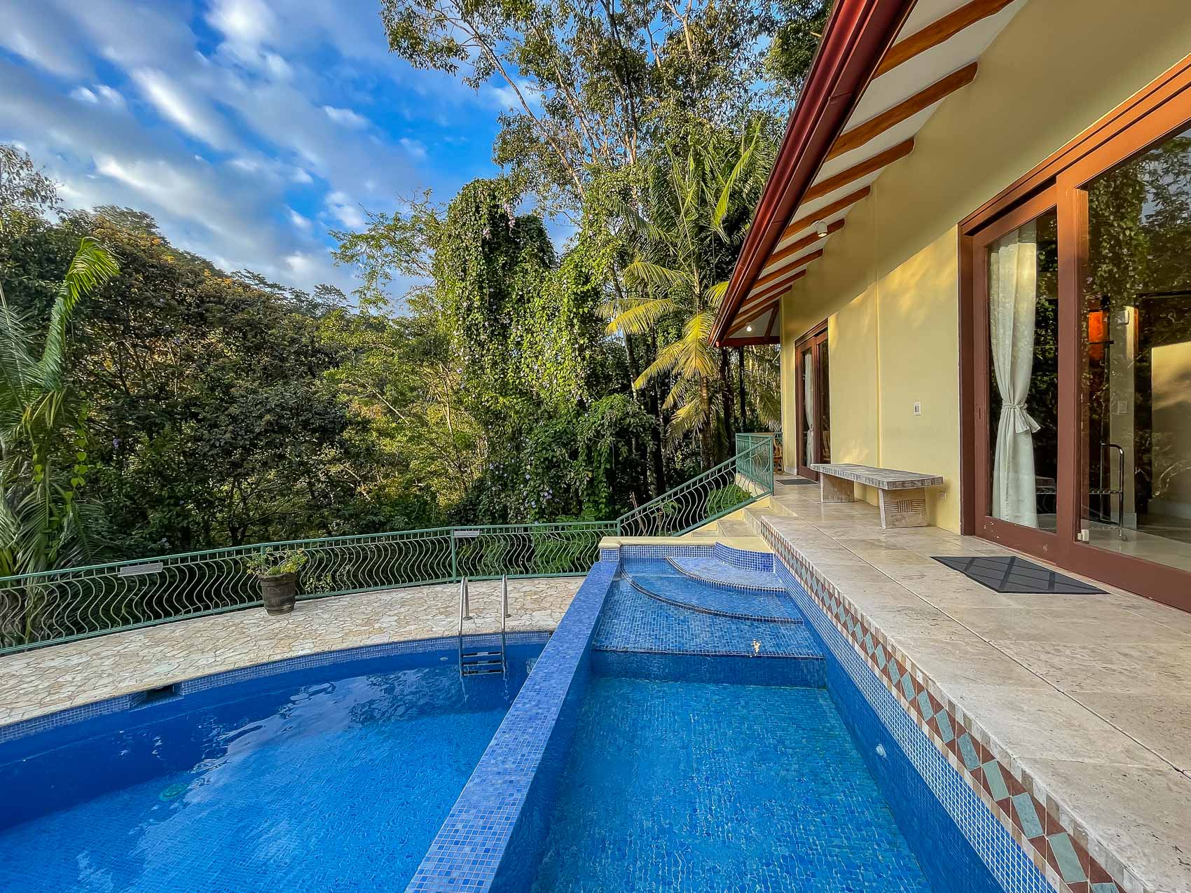 A main pool and an integrated wade pool are positioned on a tiled patio, offering direct views of lush tropical jungle under a clear sky, with glass doors opening into the villa.