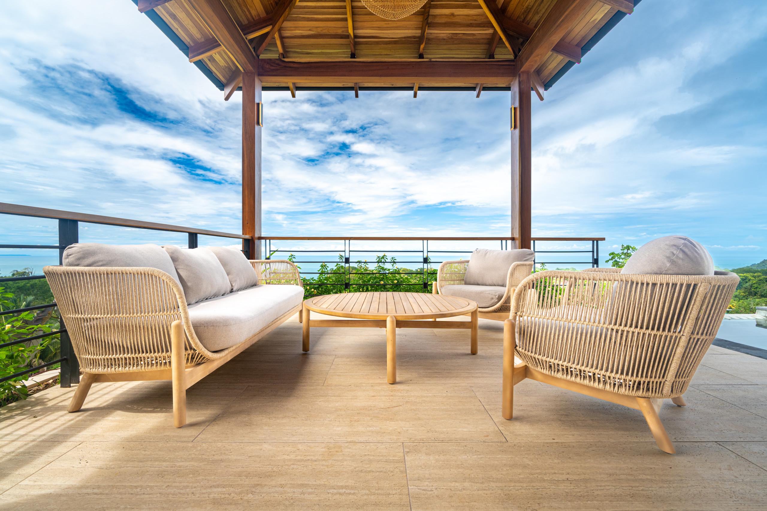 This covered terrace features a natural wood ceiling, comfortable woven seating, and panoramic ocean and jungle views, illuminated by ample natural light.