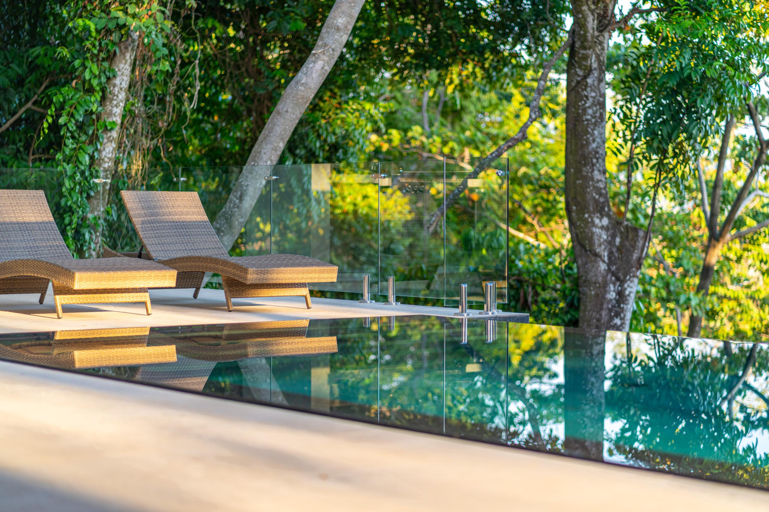 An infinity pool with a clear glass railing provides expansive views of lush tropical foliage, with two lounge chairs positioned on the adjacent terrace in warm natural light.