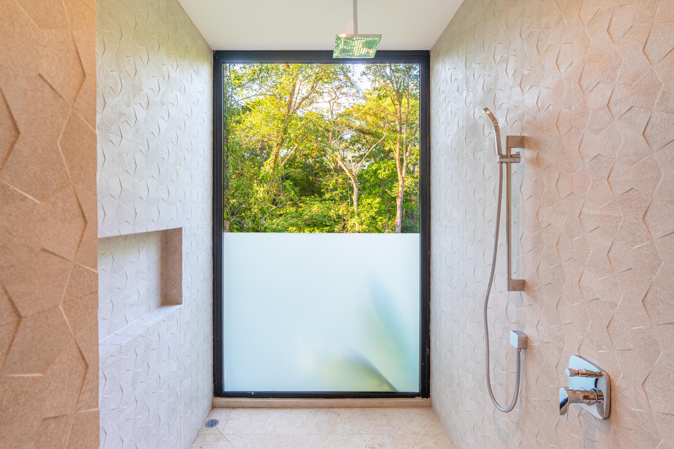 This spacious shower features textured wall tiles, a rainfall showerhead, and a large window with a frosted lower half offering private views of tropical trees.