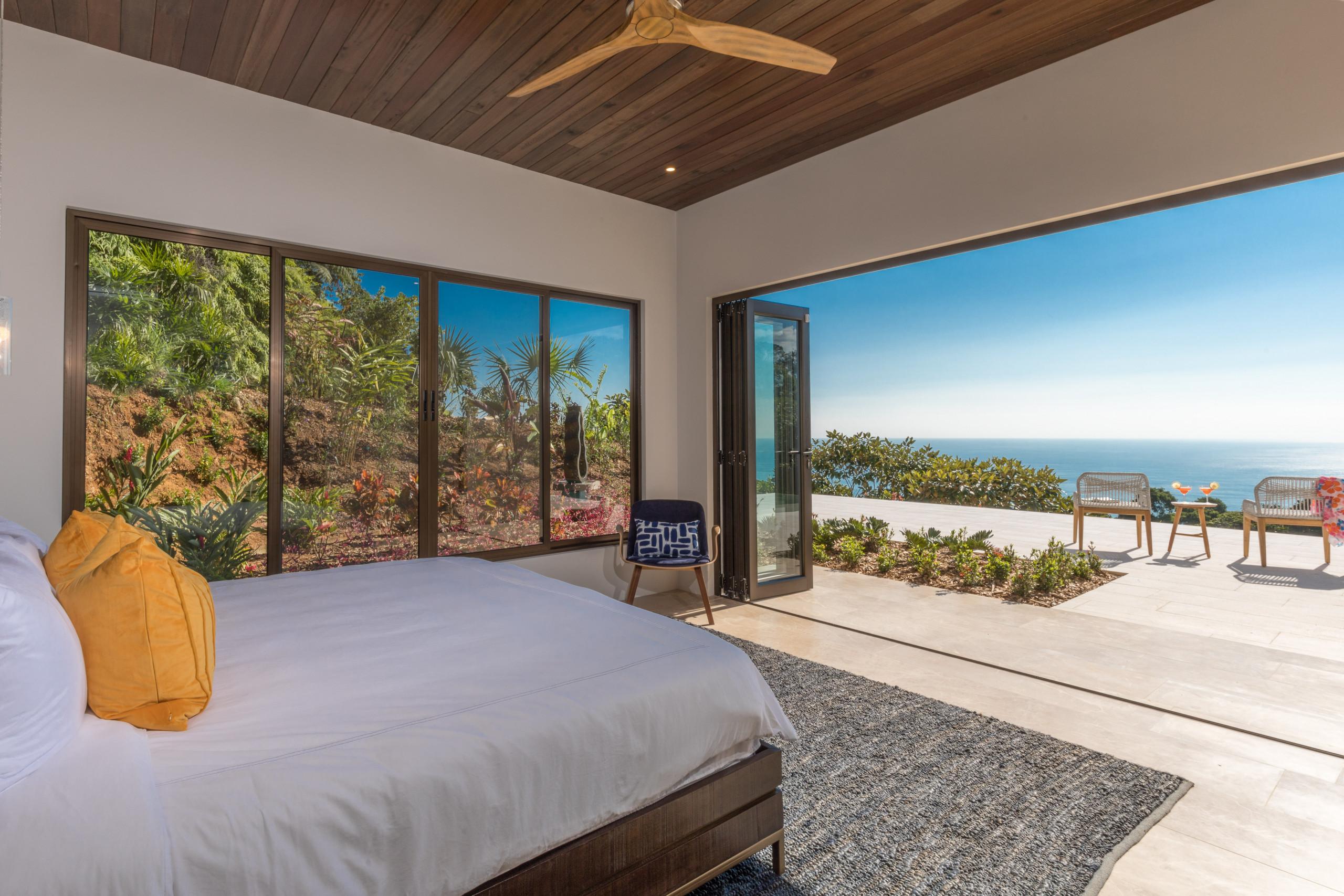 A modern bedroom with a wooden ceiling and a king-size bed offers abundant natural light, opening to a private terrace with ocean views and featuring large windows overlooking a lush jungle garden.