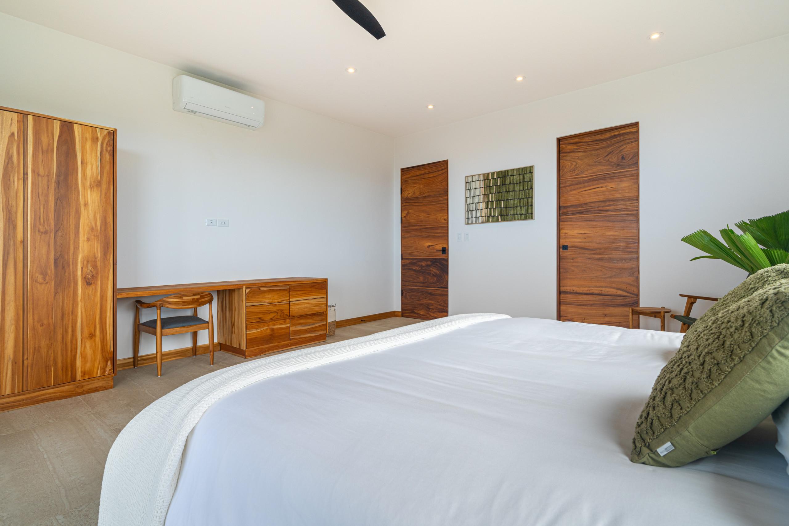 This serene bedroom features a large bed, warm-toned wooden wardrobe, desk with a chair, and two matching wooden doors set against bright white walls, complemented by a ceiling fan and air conditioning.