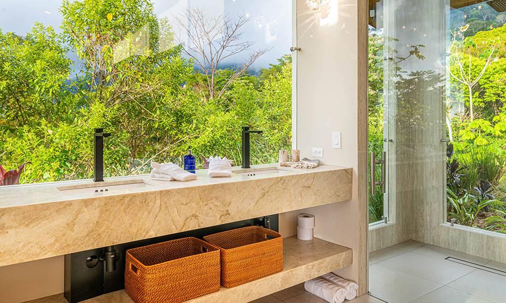 The bathroom features a stone vanity and large windows offering views of the surrounding jungle and filling the space with natural light.