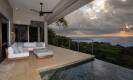 A covered terrace with a lounge area and infinity pool offers expansive ocean views under a cloudy sunset sky.