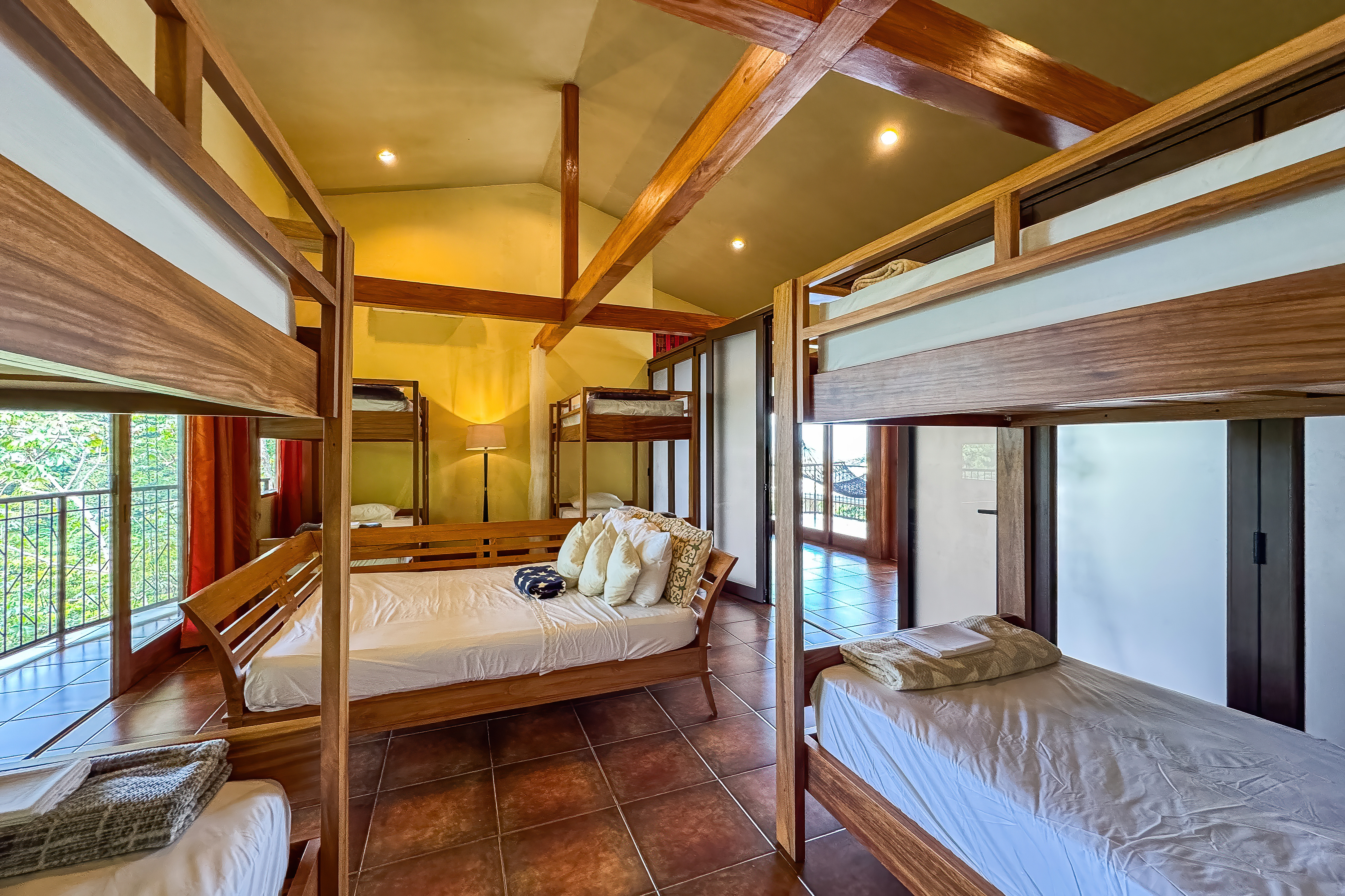 A bright, shared bedroom with high vaulted ceilings, exposed wood beams, multiple bunk beds, and a double bed, provides views of the rainforest and access to an adjacent outdoor space.