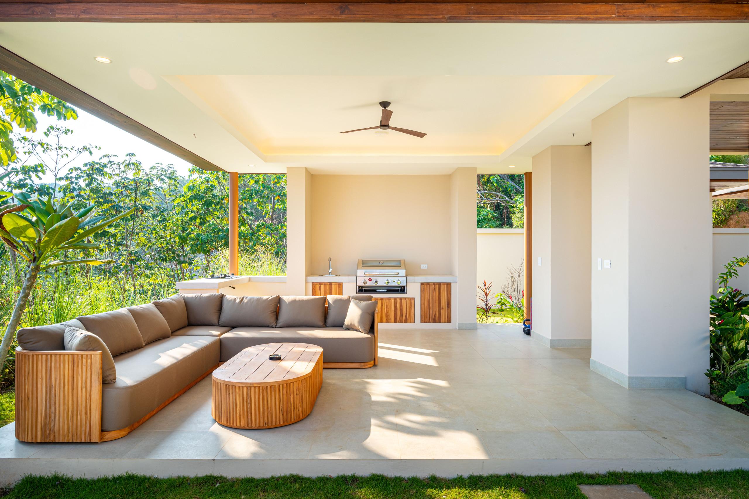 A shaded outdoor terrace with Balinese-inspired wooden lounge seating and a built-in kitchen area provides a seamless connection to the natural light and surrounding rainforest.