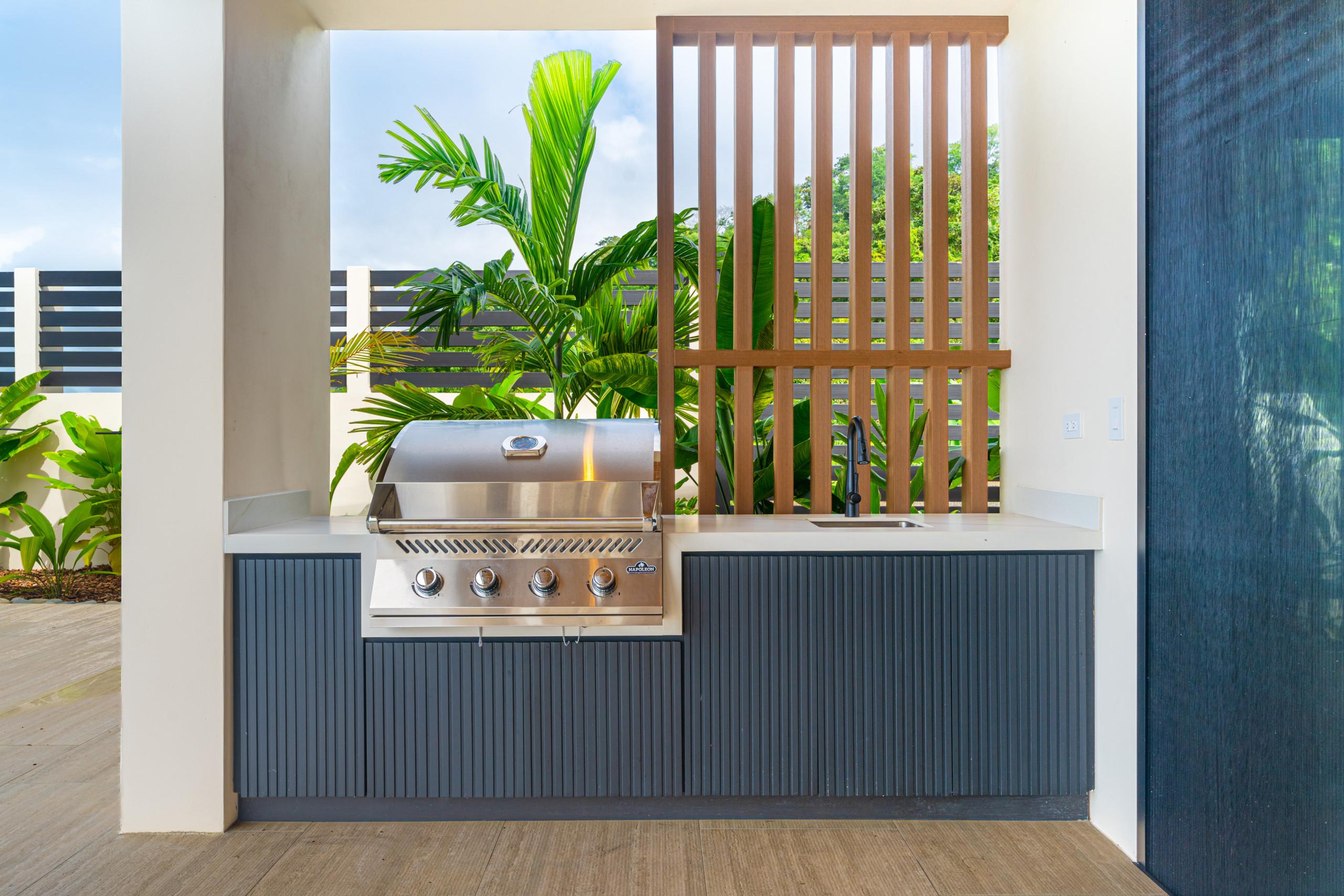 A covered outdoor kitchen features a built-in stainless steel grill and sink, complemented by dark ribbed cabinetry and tropical greenery visible through a wooden slat screen.